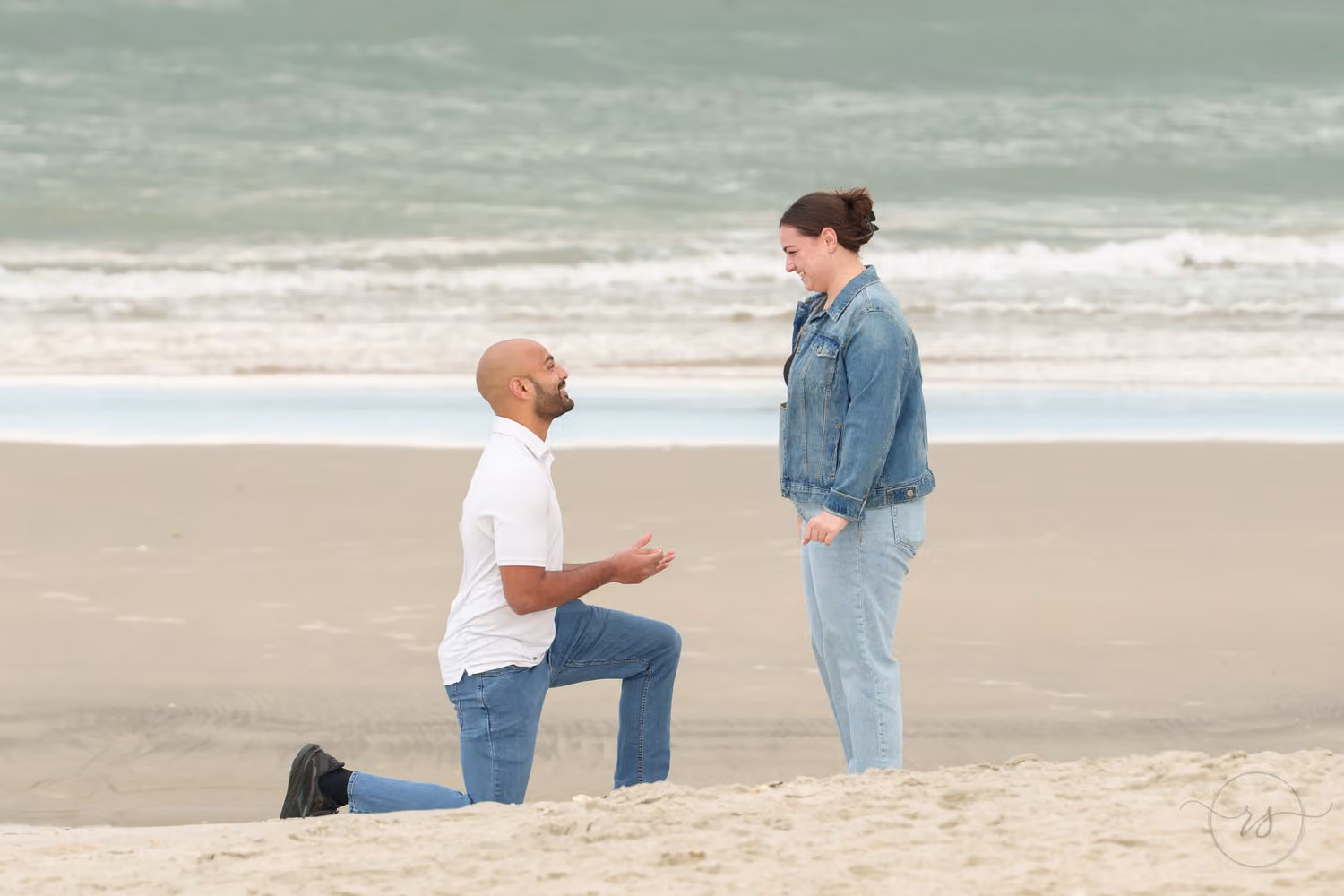 Man kneeling on the sand proposing to a smiling woman in a denim jacket by the waves at Huntington Beach State Park