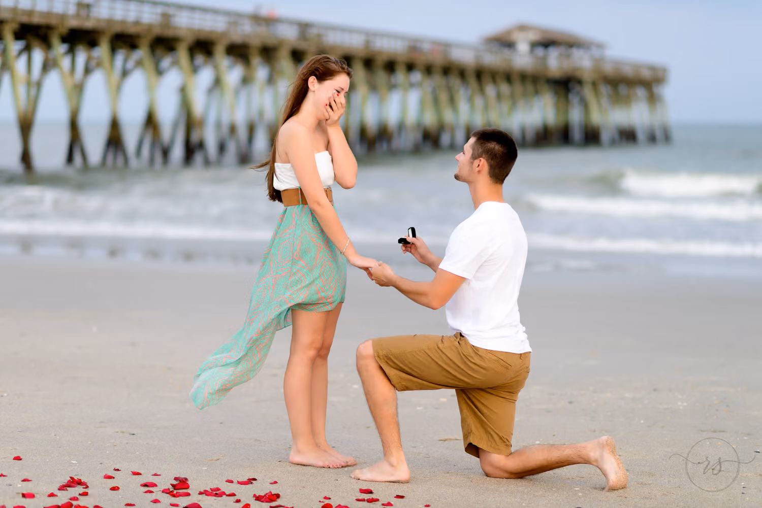 Man on one knee proposing on rose petals in the sand as she covers her mouth, with the pier behind them in Myrtle Beach