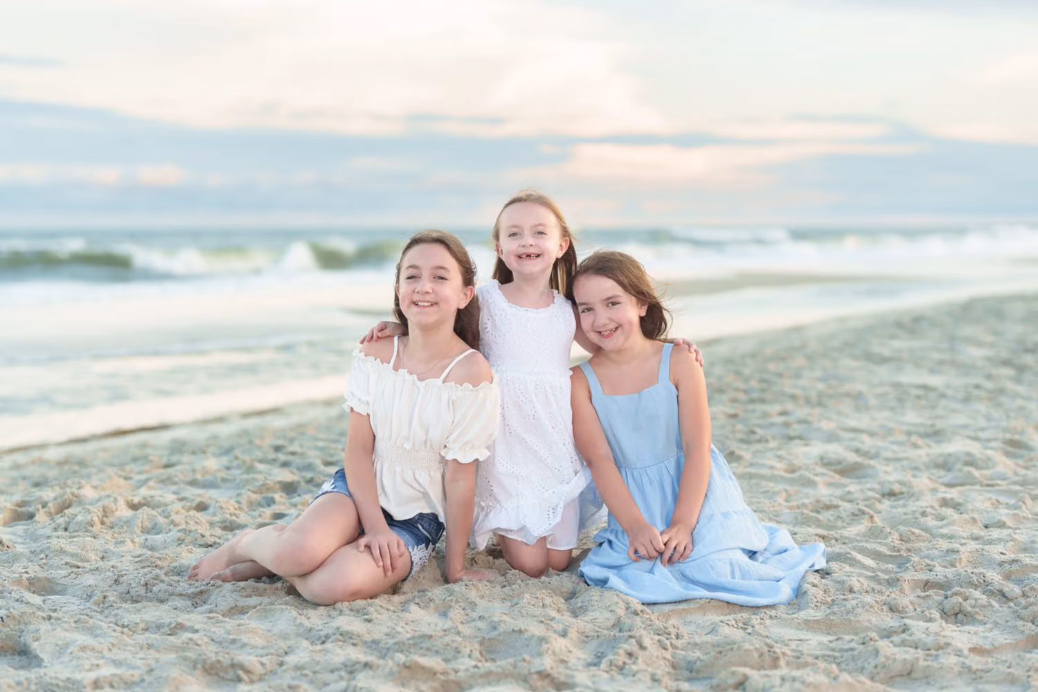 Three little sisters sitting together in the sand with the ocean behind them at Huntington Beach State Park