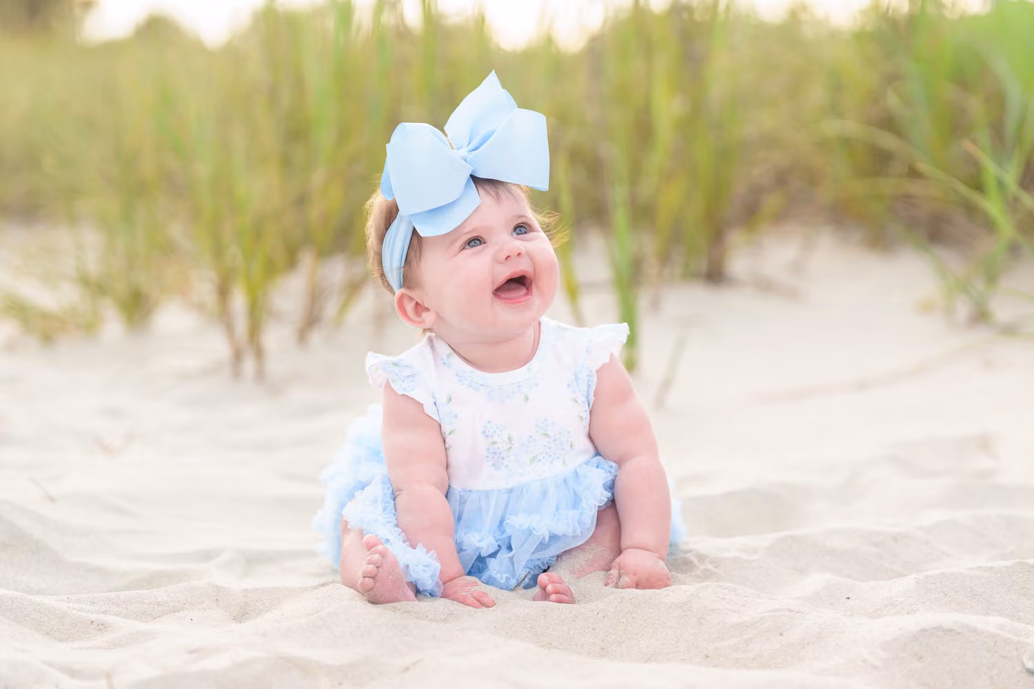Laughing baby girl with a big blue bow sitting in the sand in front of dune grass in North Myrtle Beach