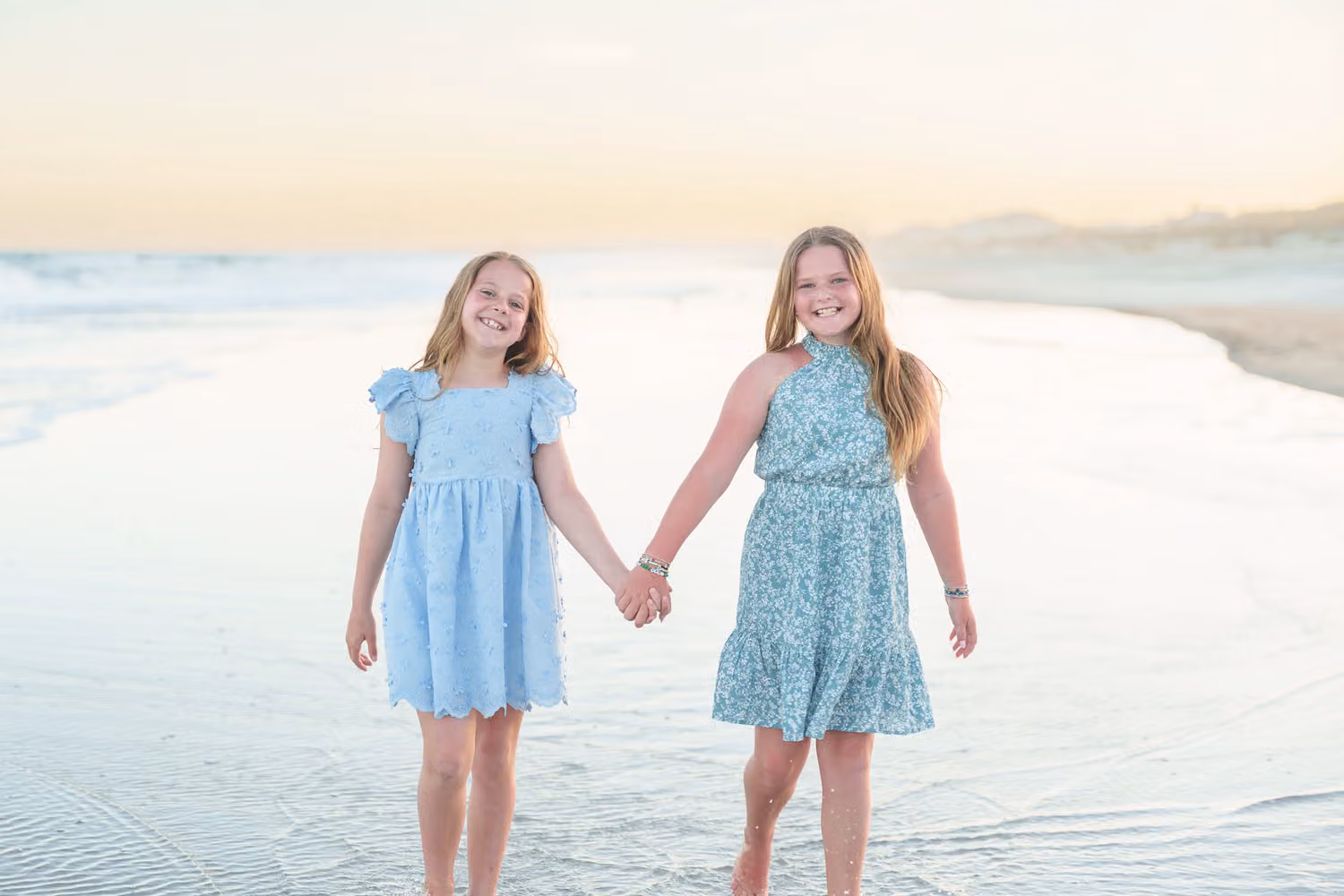 Two girls in blue dresses holding hands and walking along the wet sand at sunset at Huntington Beach State Park
