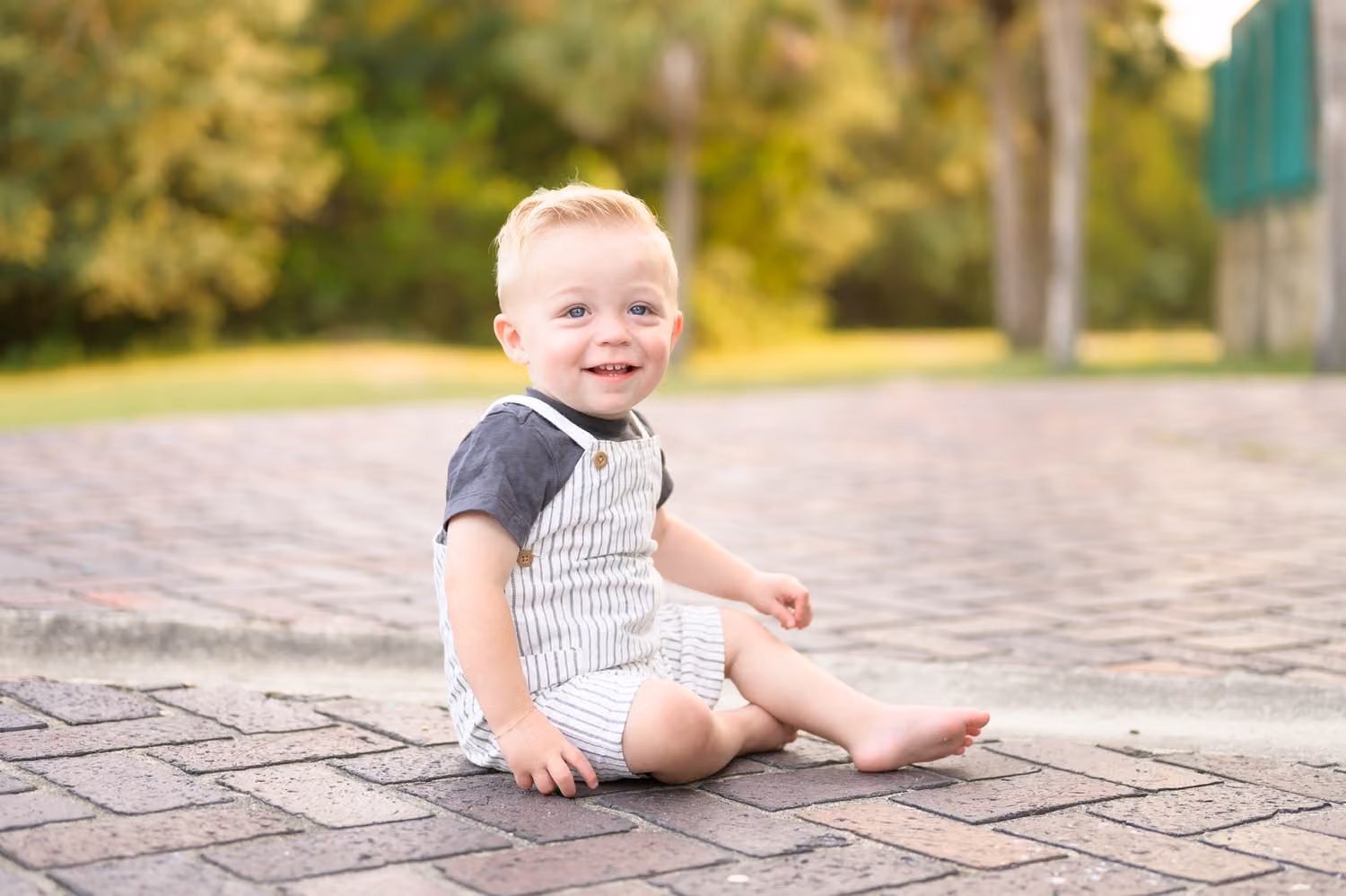 Smiling baby boy in striped overalls sitting on the brick pavers behind the castle at Huntington Beach State Park