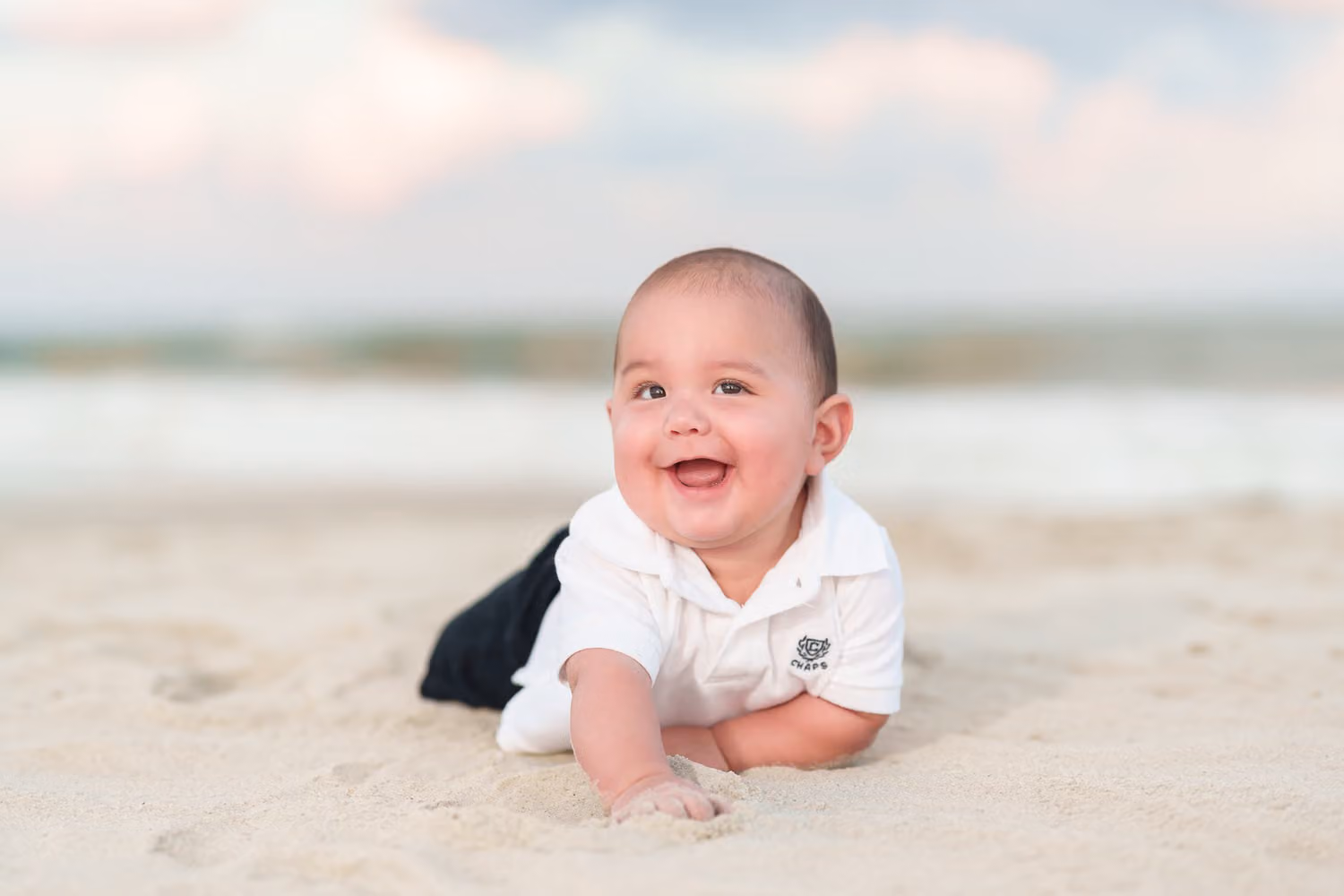 Newly adopted baby boy on his tummy in the sand grinning with the ocean behind him at Huntington Beach State Park