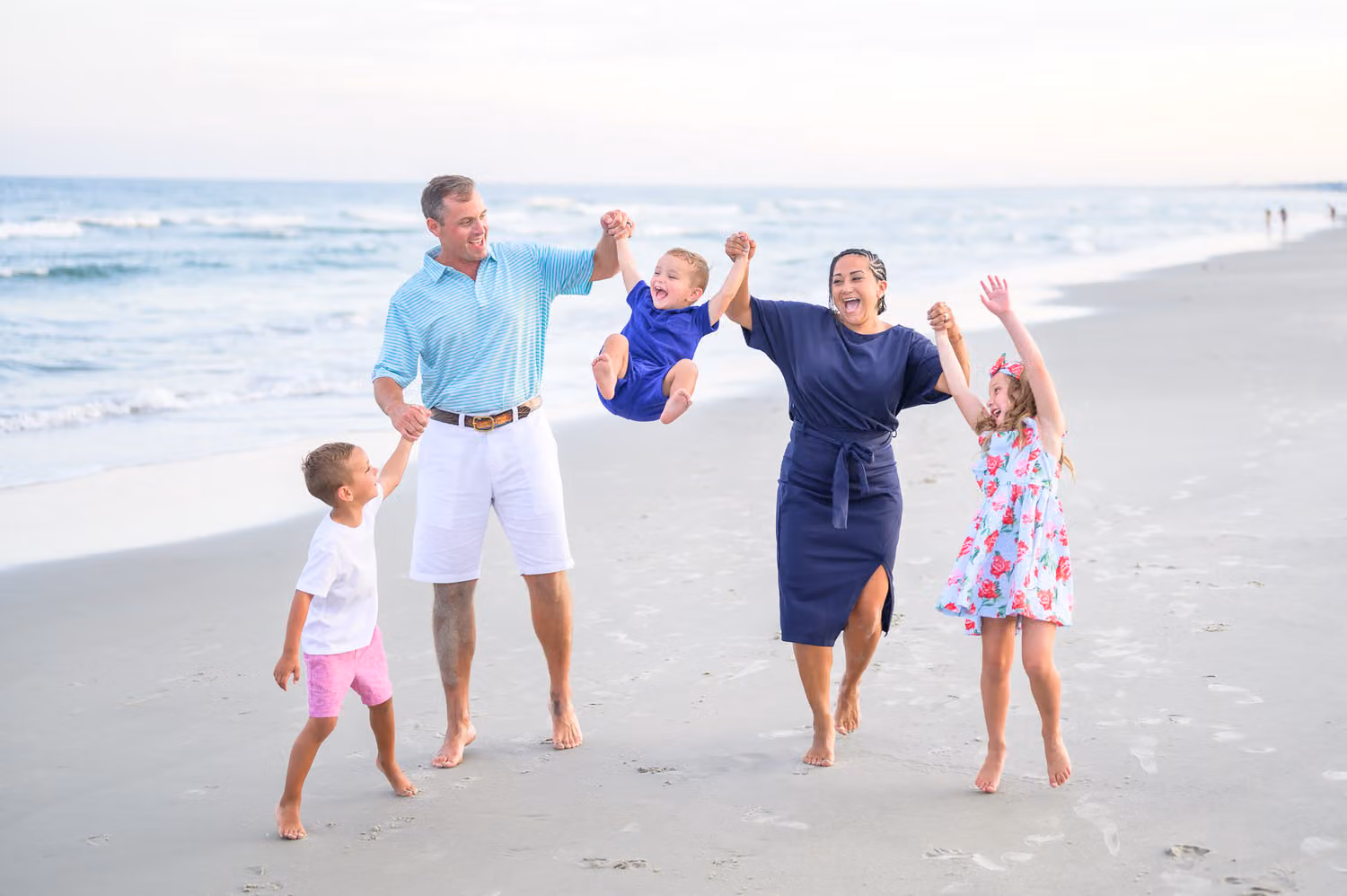 Mom and dad swinging their toddler between them as two kids dance alongside on the beach at Huntington Beach State Park