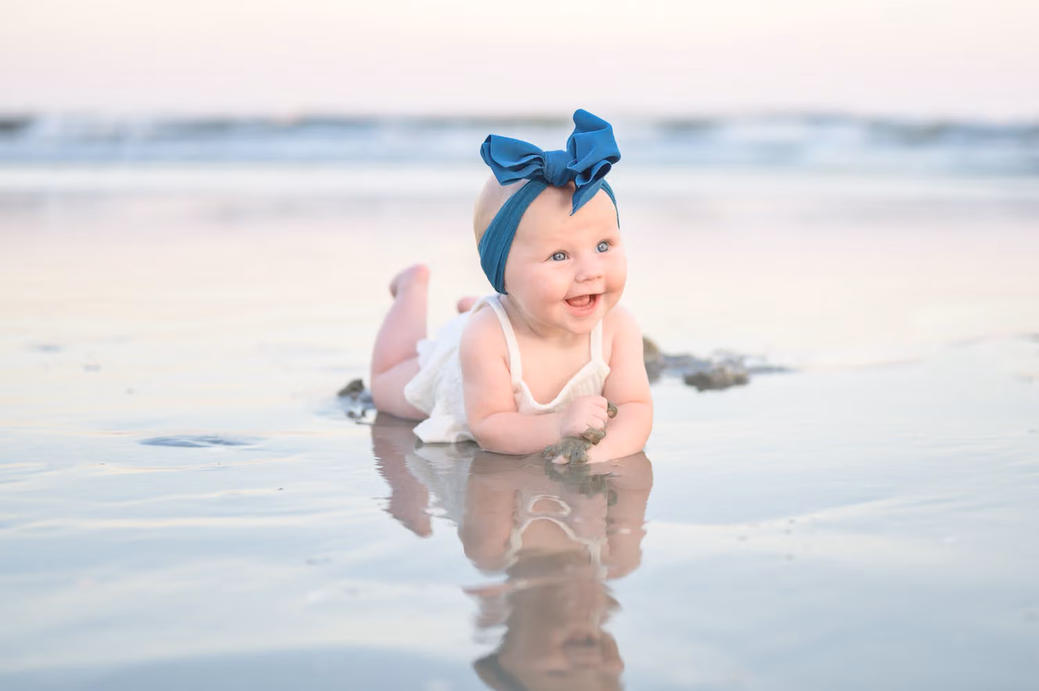 Smiling baby girl in a big blue bow lying on her tummy in shallow water with her reflection in North Myrtle Beach