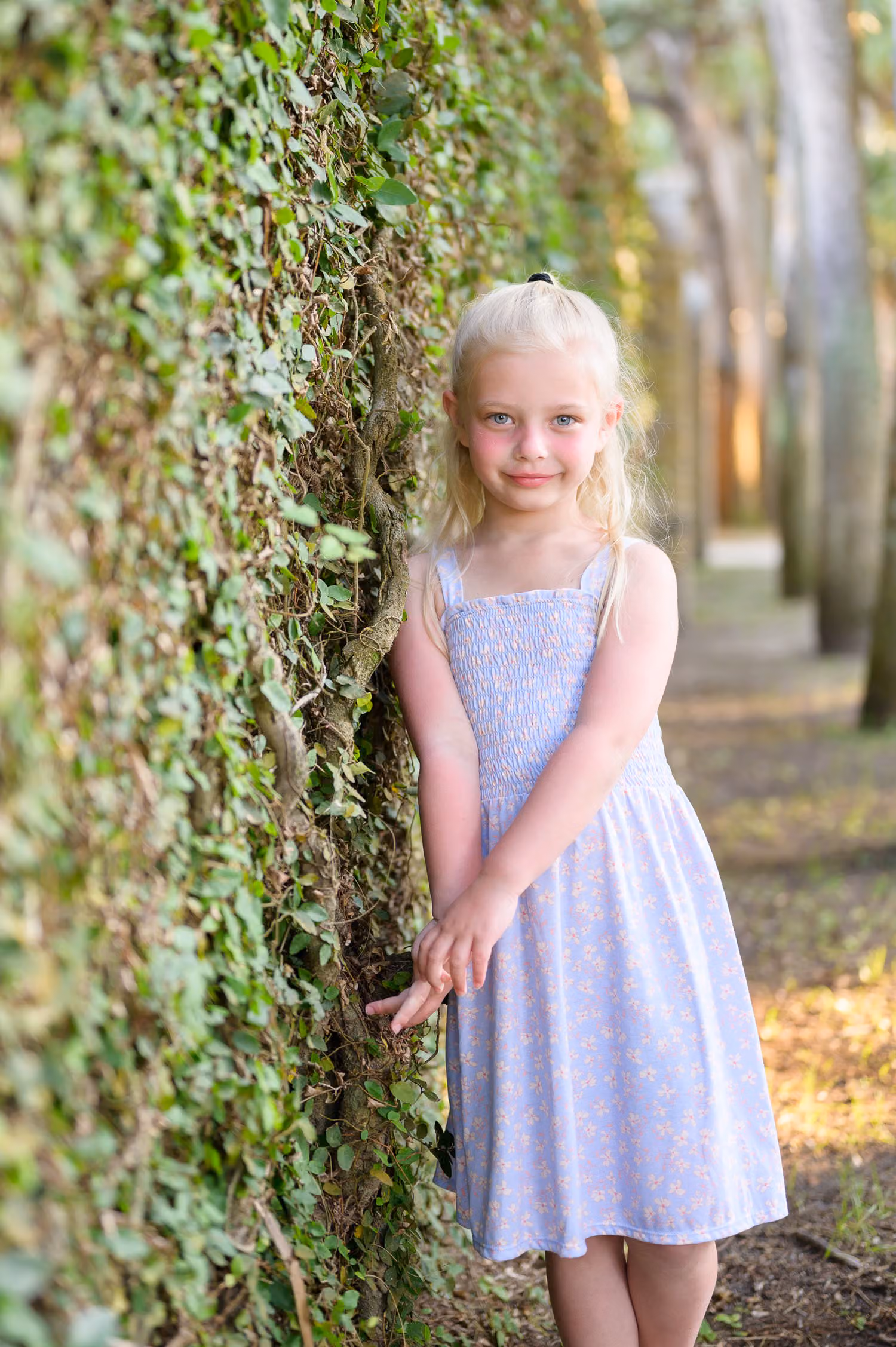 Blonde little girl in a lavender sundress leaning against the ivy wall in soft light at Huntington Beach State Park