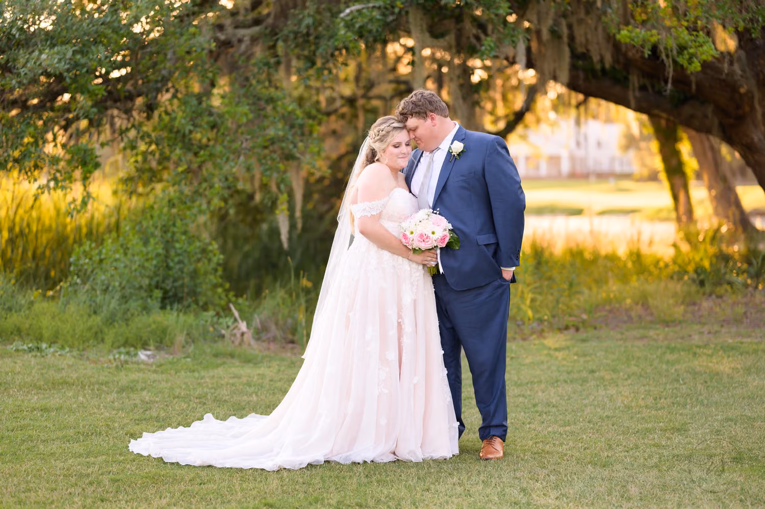 Bride holding her bouquet and groom in a blue suit embracing under oak branches on the golf course at Pawleys Plantation