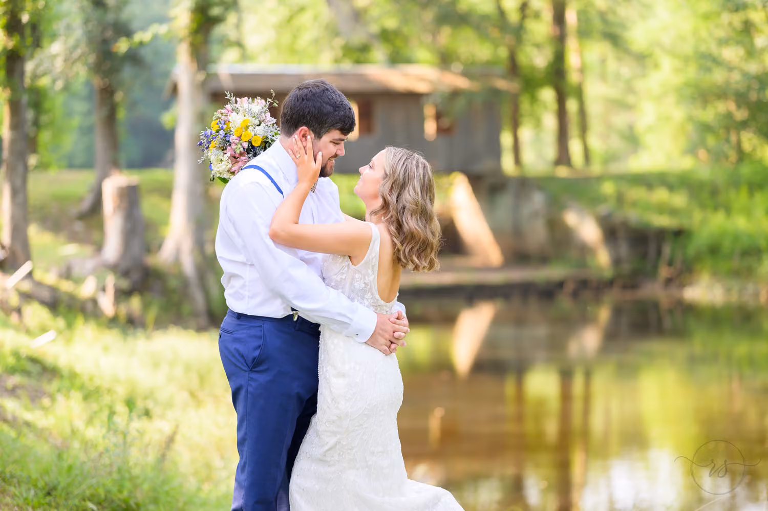 Bride and groom holding each other close by a pond with an old wooden bridge behind them at Pine Log Plantation