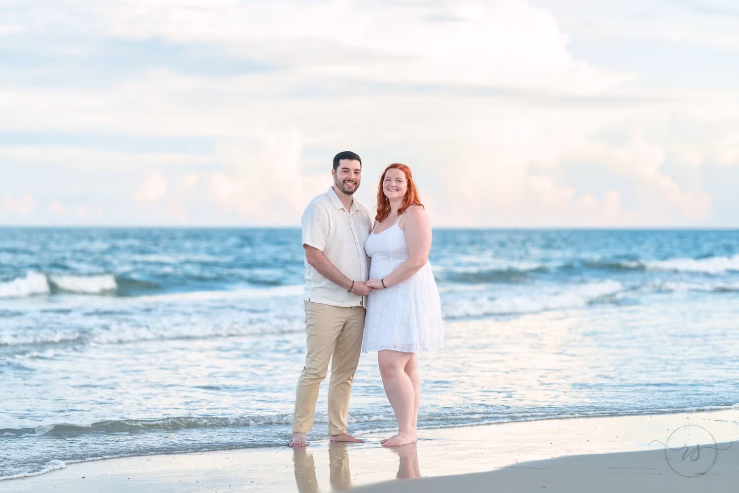Engaged couple in light clothes holding hands barefoot at the water's edge with waves and pastel clouds behind them