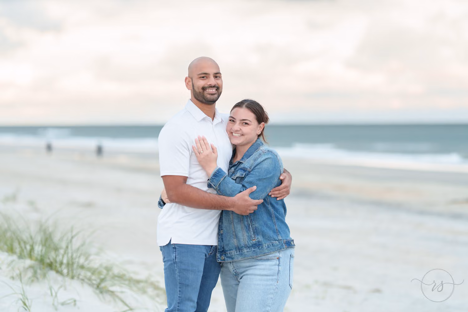 Smiling couple hugging on the beach by the dune grass, her ring hand on his chest, at Huntington Beach State Park