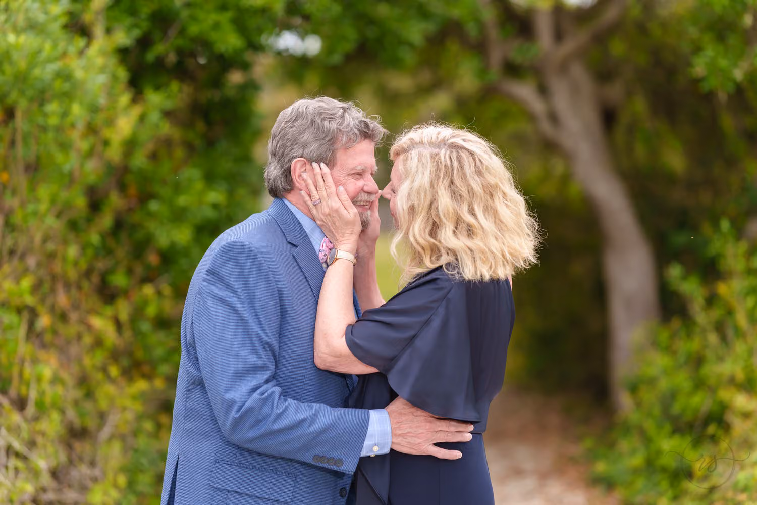 Older couple holding each other's faces lovingly on a leafy path at Huntington Beach State Park