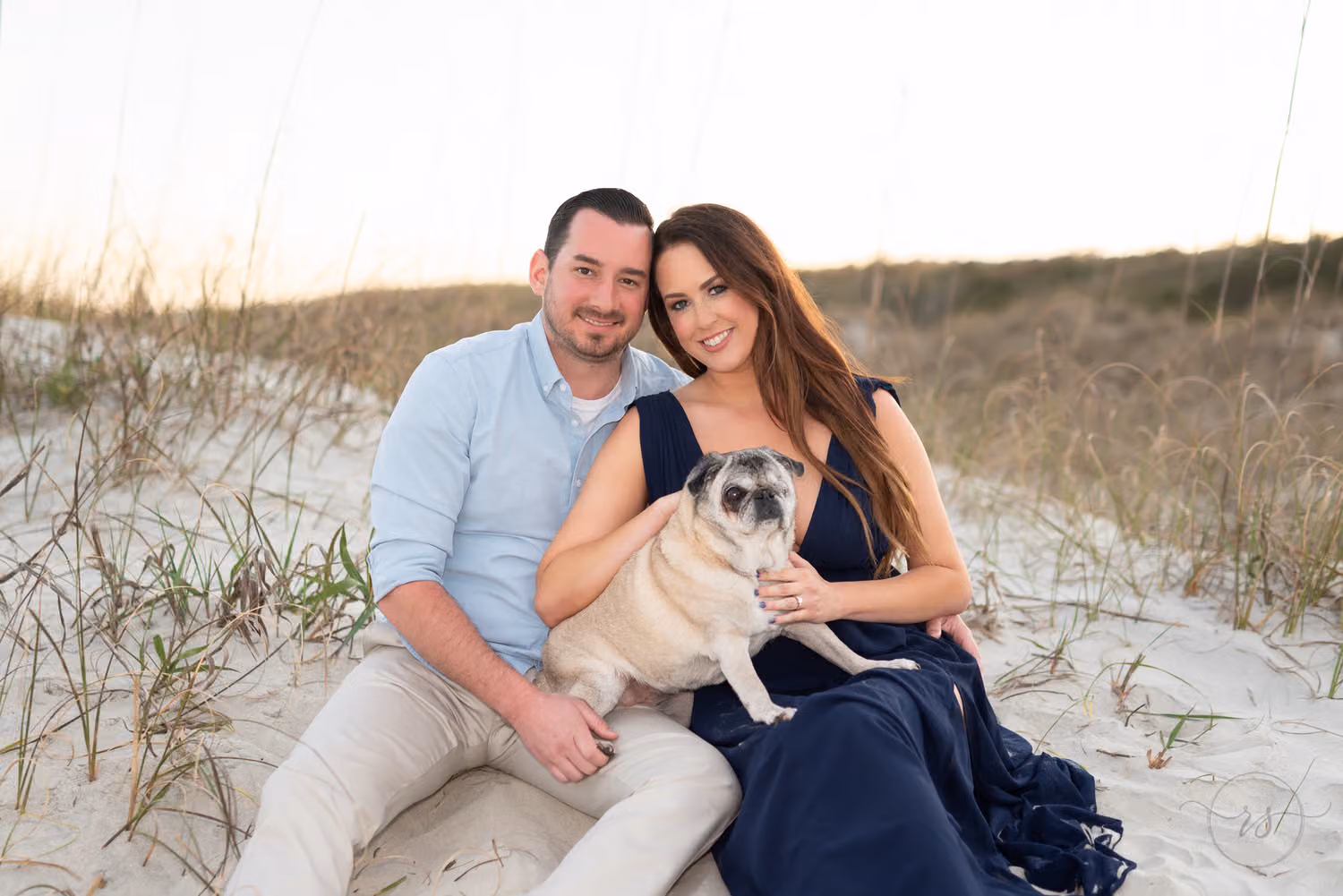 Couple sitting in the sand in front of the dunes holding their pug at Huntington Beach State Park