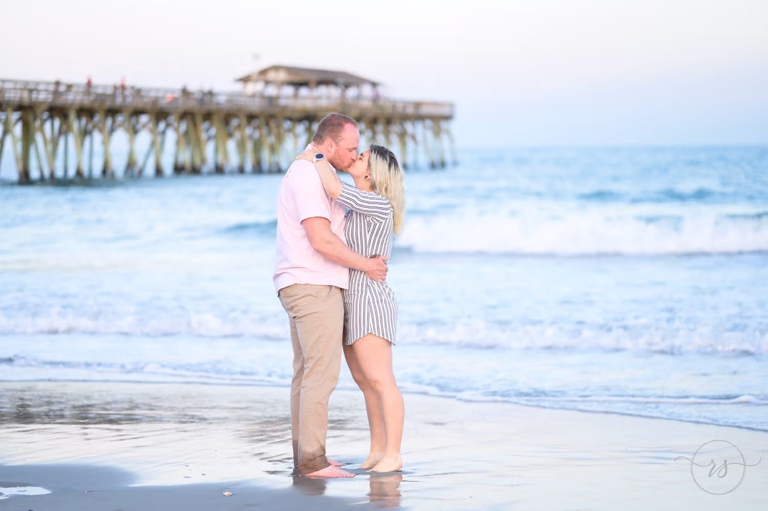 Couple kissing at the water's edge with the fishing pier stretching out behind them at Myrtle Beach State Park
