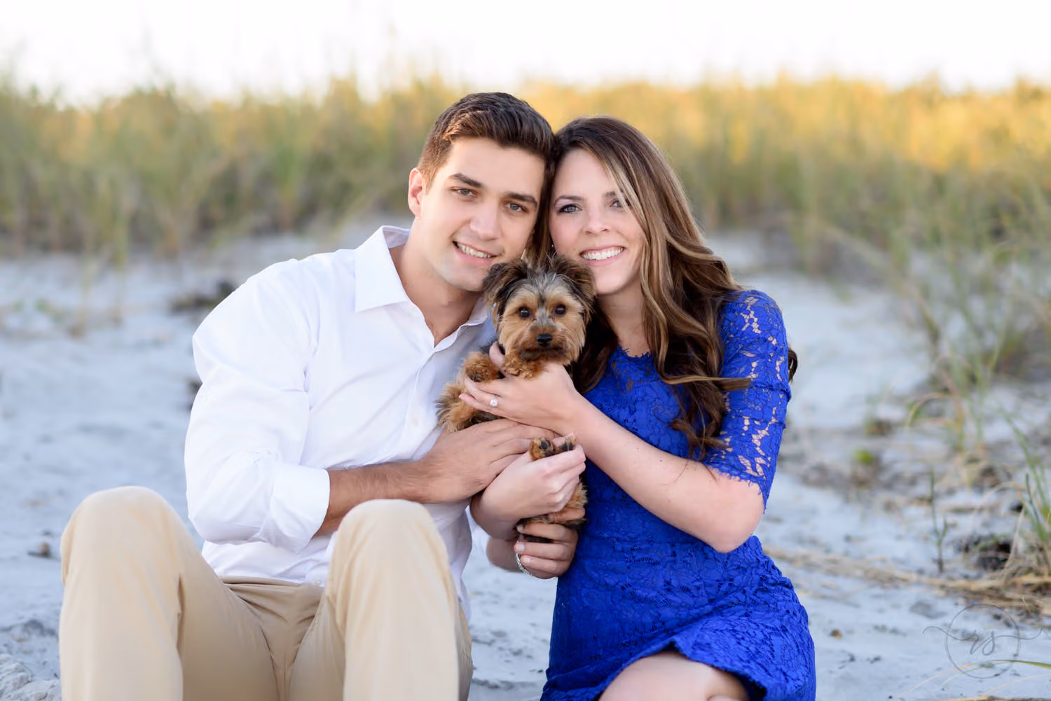 Couple sitting in front of the dunes holding their small puppy between them at Myrtle Beach State Park