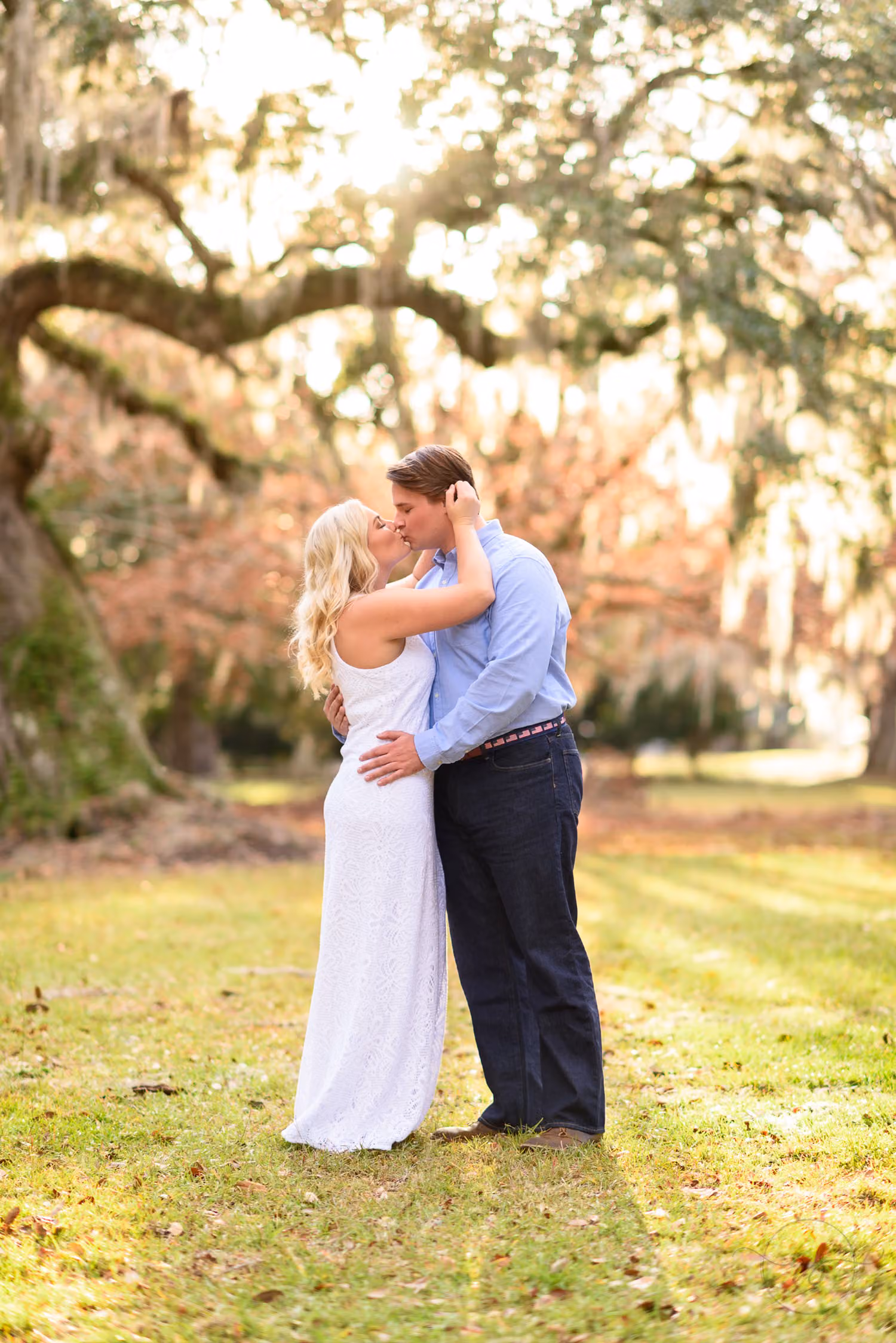 Couple in white and blue kissing on the lawn under huge oak trees with Spanish moss at Litchfield Plantation