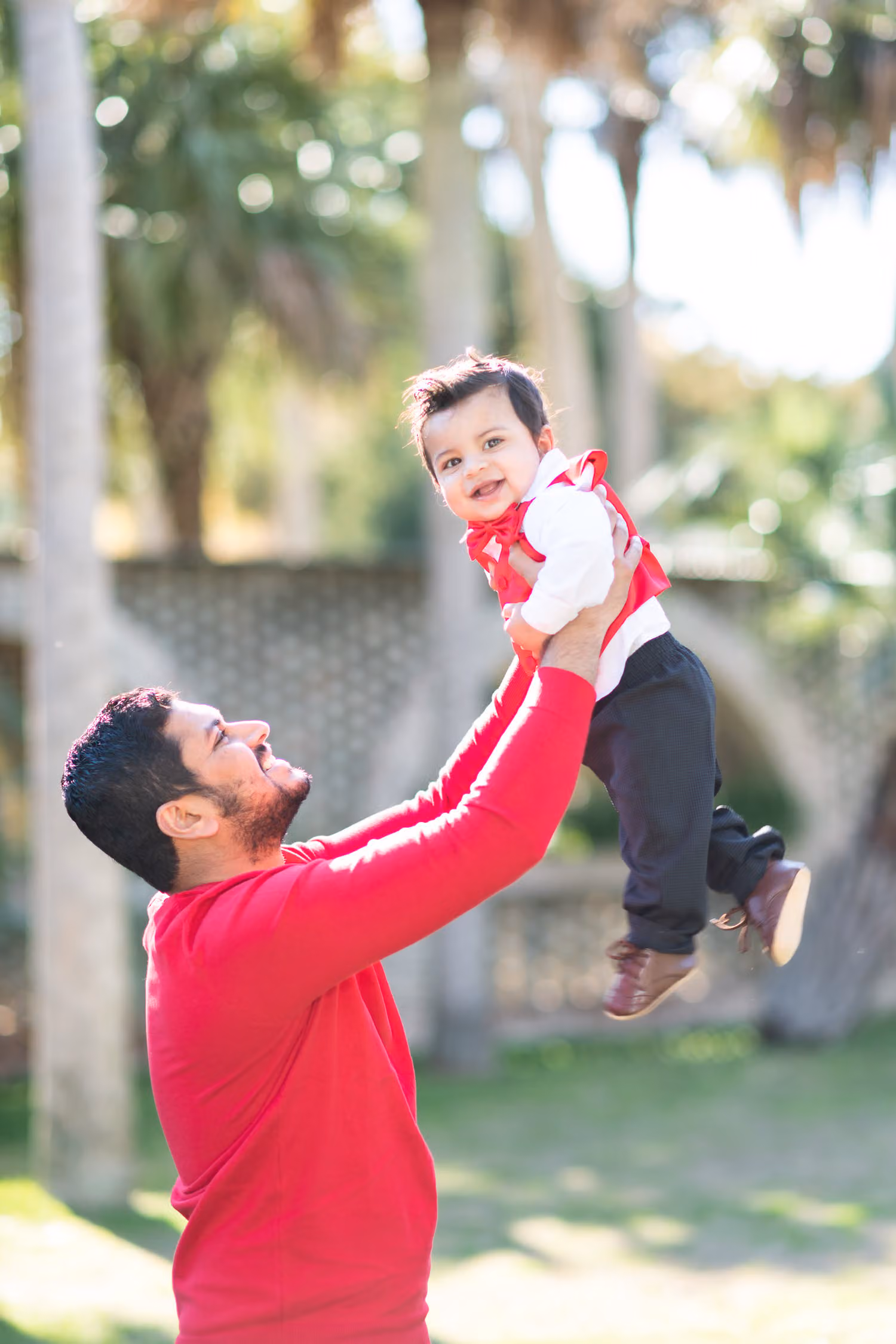 Dad in a red sweater lifting his laughing baby boy into the air in front of palm trees at Atalaya Castle