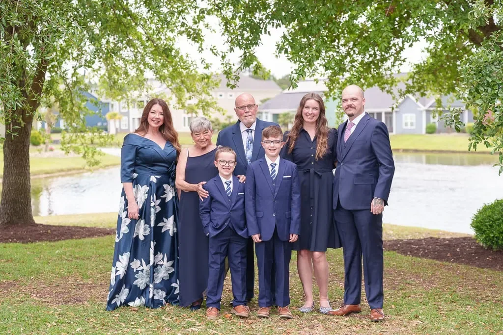 Family in navy formal wear standing together on a lawn beside a pond