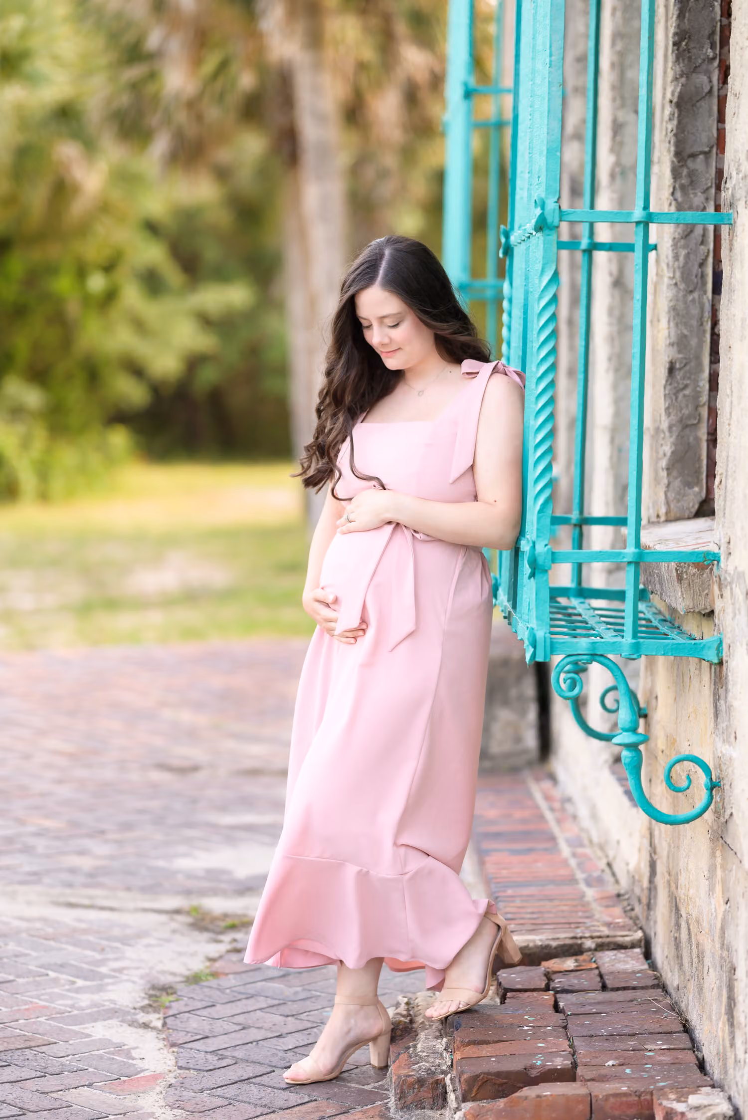 Expectant mother in a pink dress leaning by a teal barred window at Atalaya Castle, Huntington Beach State Park