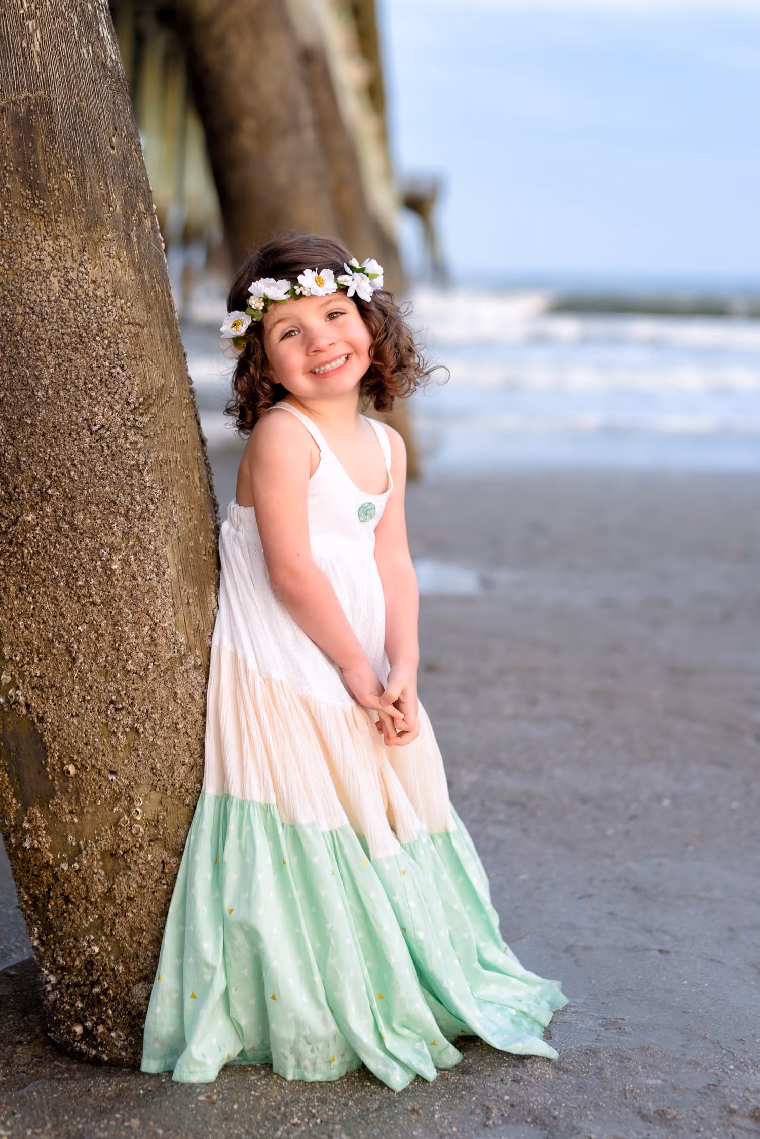Little girl in a flower crown and long dress leaning against a pier piling at Myrtle Beach State Park
