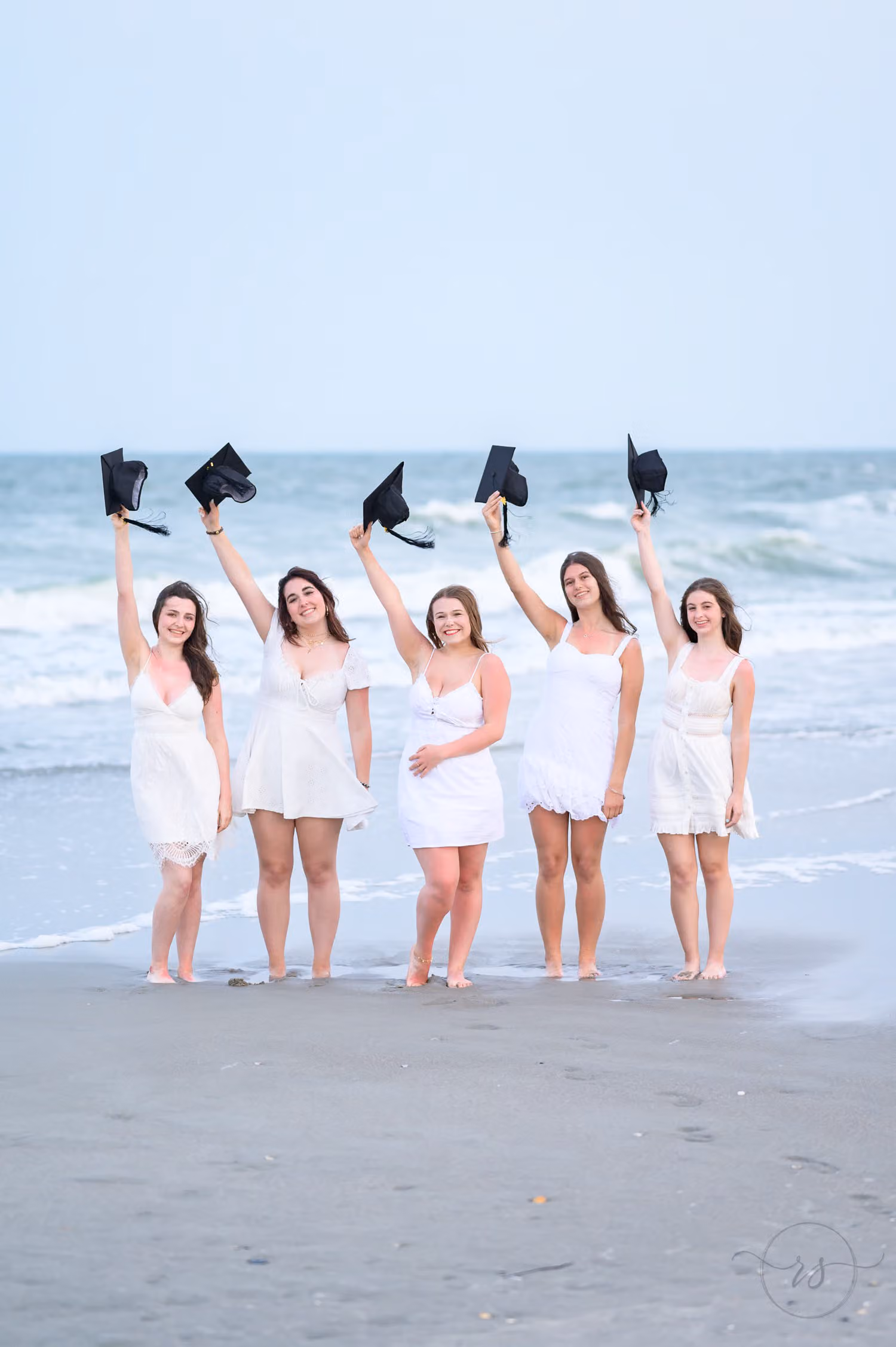 Five seniors in white dresses raising their graduation caps on the beach at Myrtle Beach State Park
