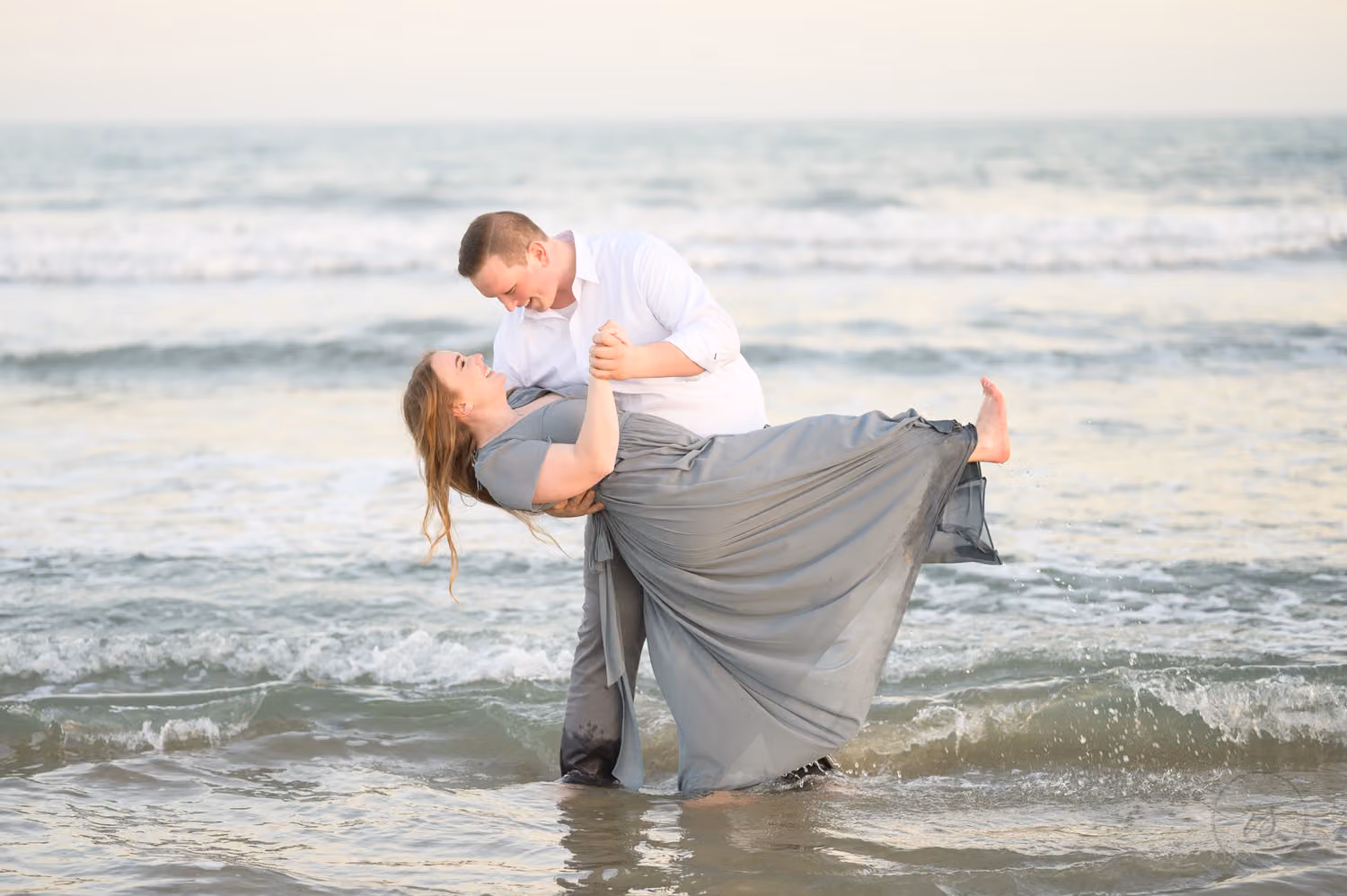 Man dipping his fiancée in a long gray dress in the shallow surf at Huntington Beach State Park