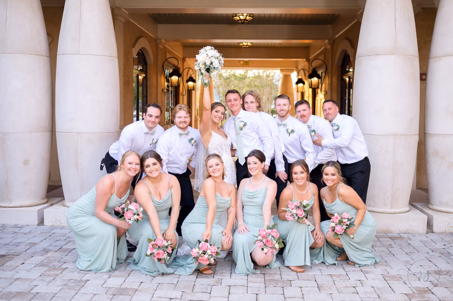 Bridesmaids in sage green crouching in front of laughing groomsmen between the courtyard columns at 21 Main Events