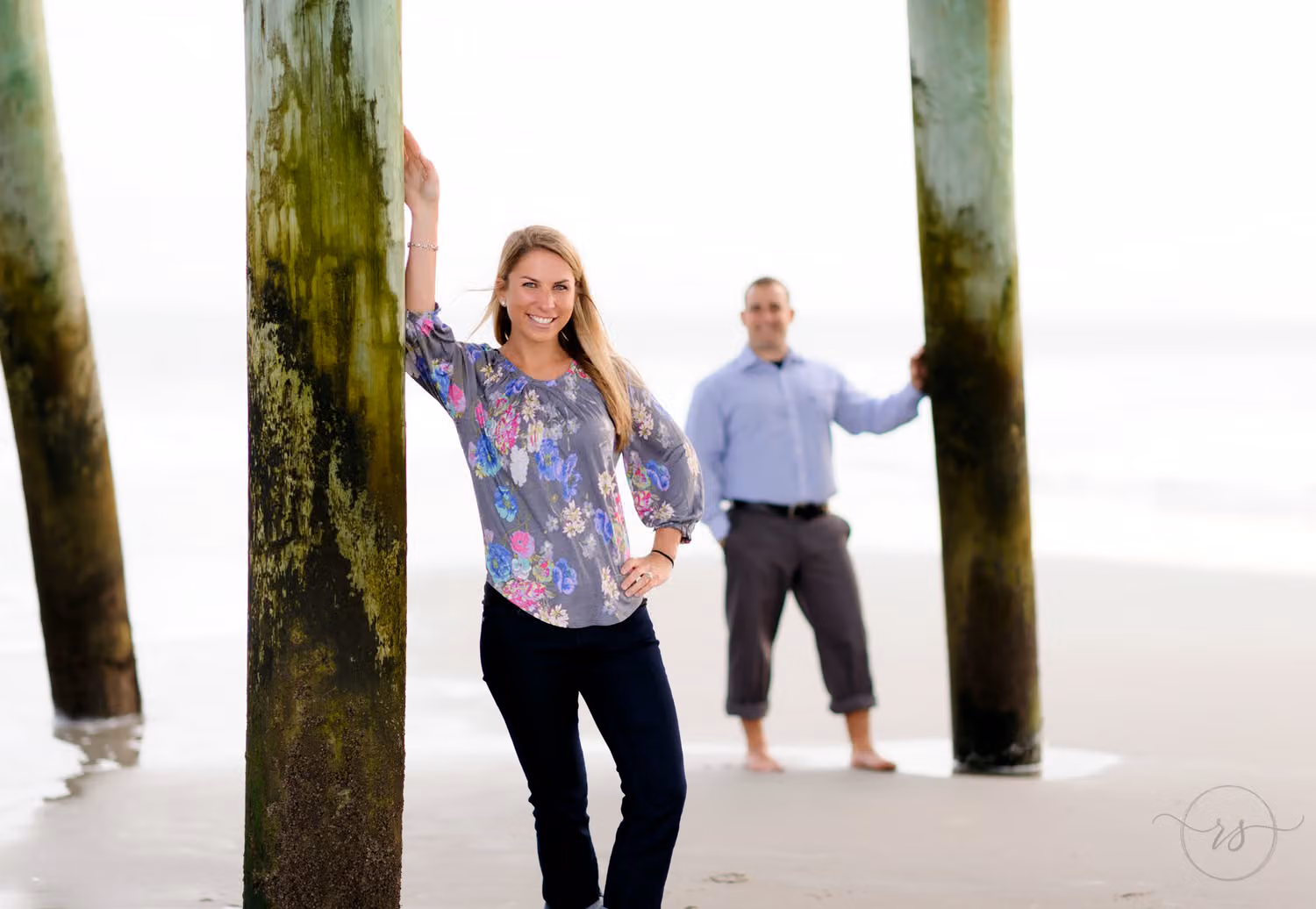 Woman leaning on a pier piling in focus while her fiancé waits behind her on the beach at Myrtle Beach State Park