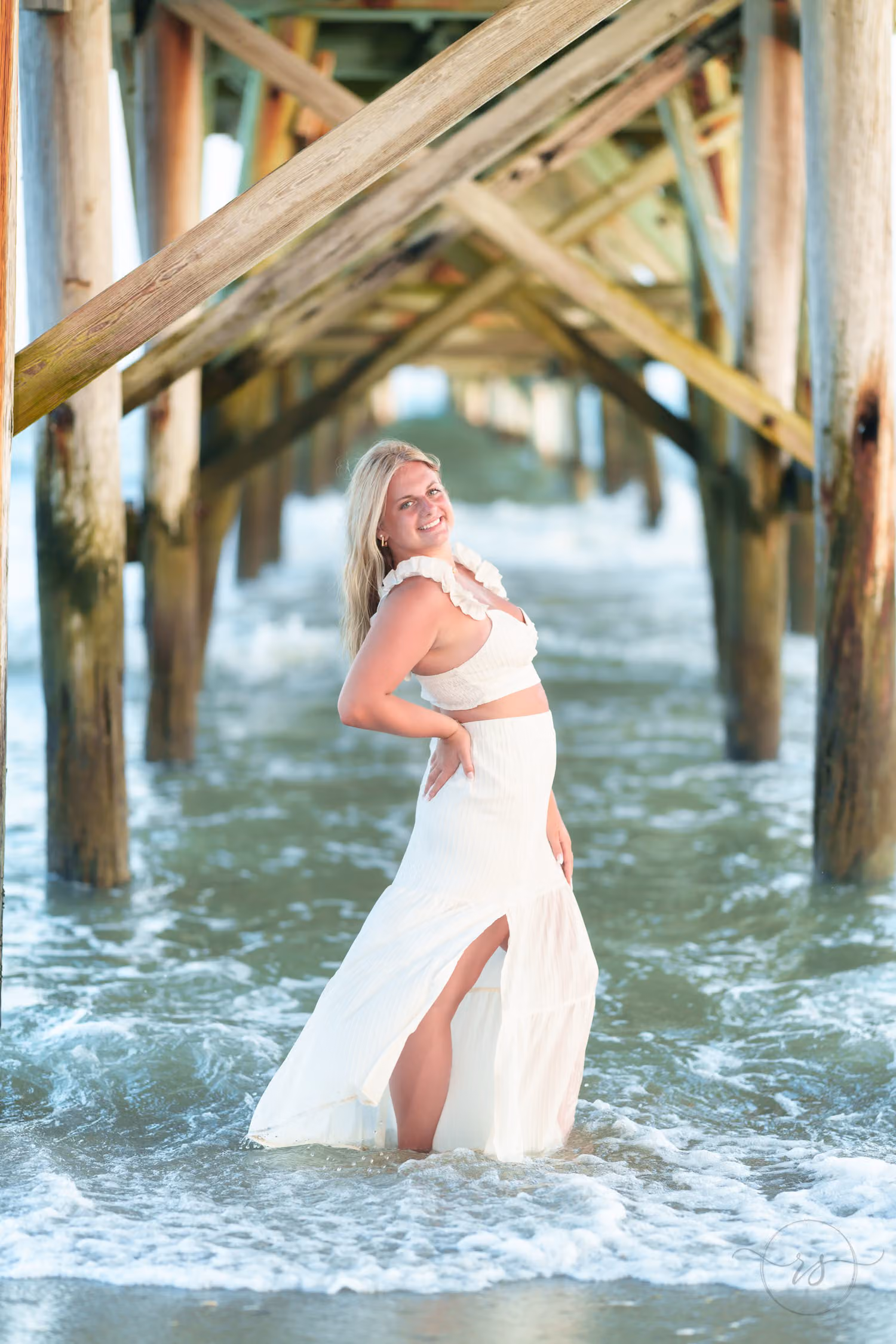 Senior girl in a white top and long white skirt standing in the water beneath the pier's wooden beams in North Myrtle Beach