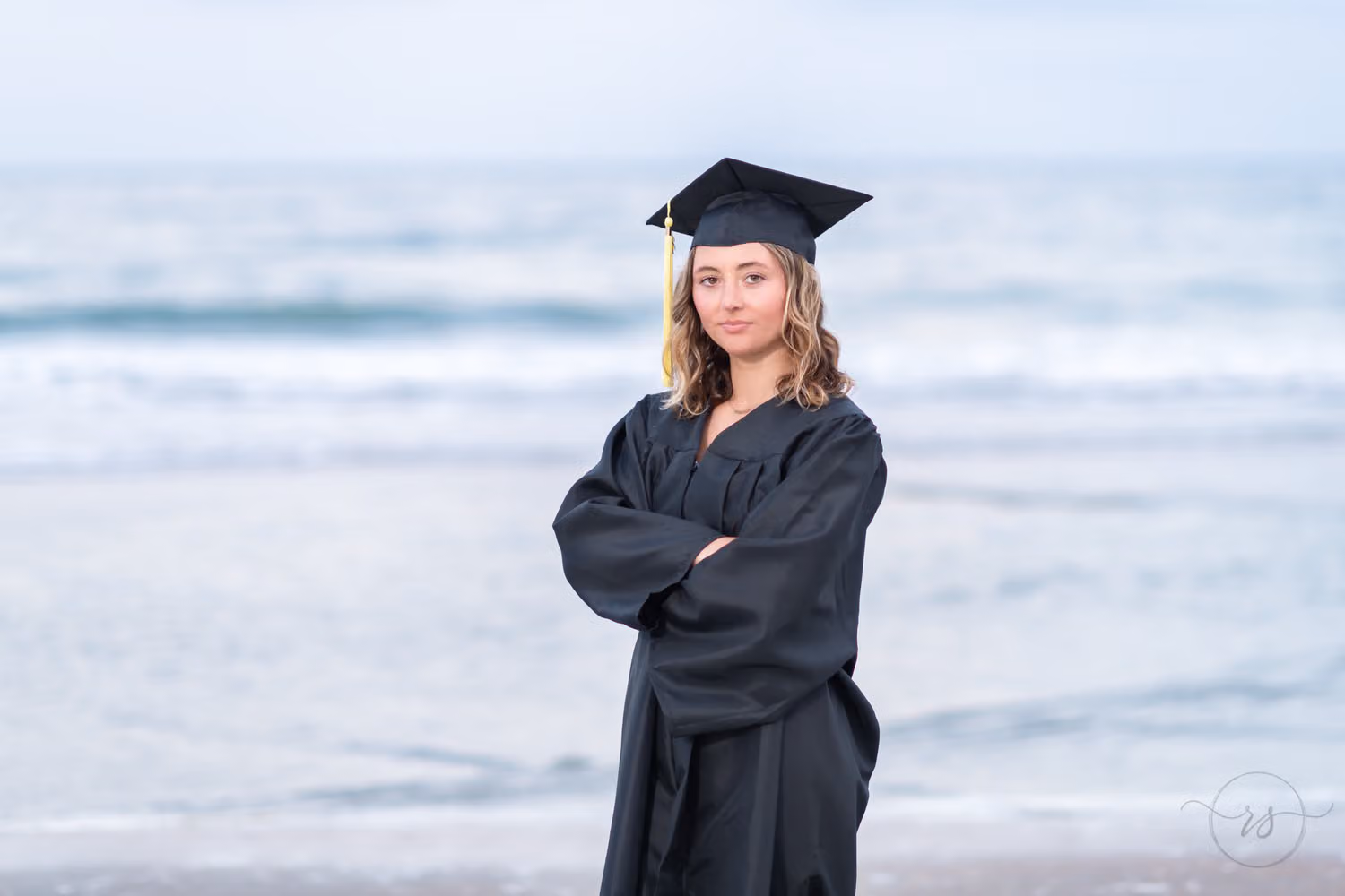 Graduate in a black cap and gown with her arms crossed in front of the ocean at Huntington Beach State Park