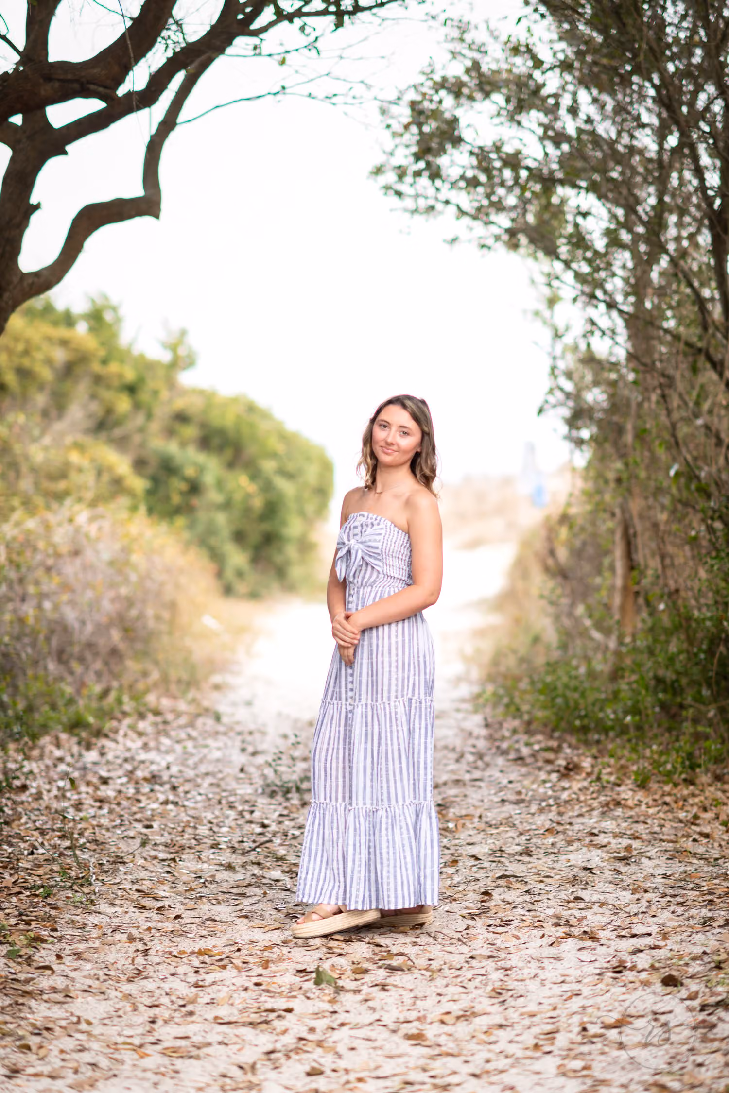 Senior in a long striped dress standing on a leafy path under arching trees at Huntington Beach State Park
