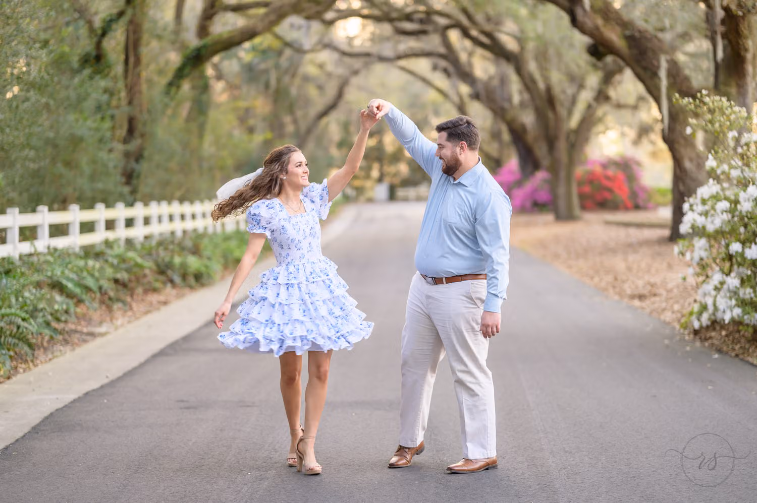 Man spinning his fiancée in a blue floral dress on a drive lined with mossy oaks and azaleas at Caledonia Golf and Fish Club