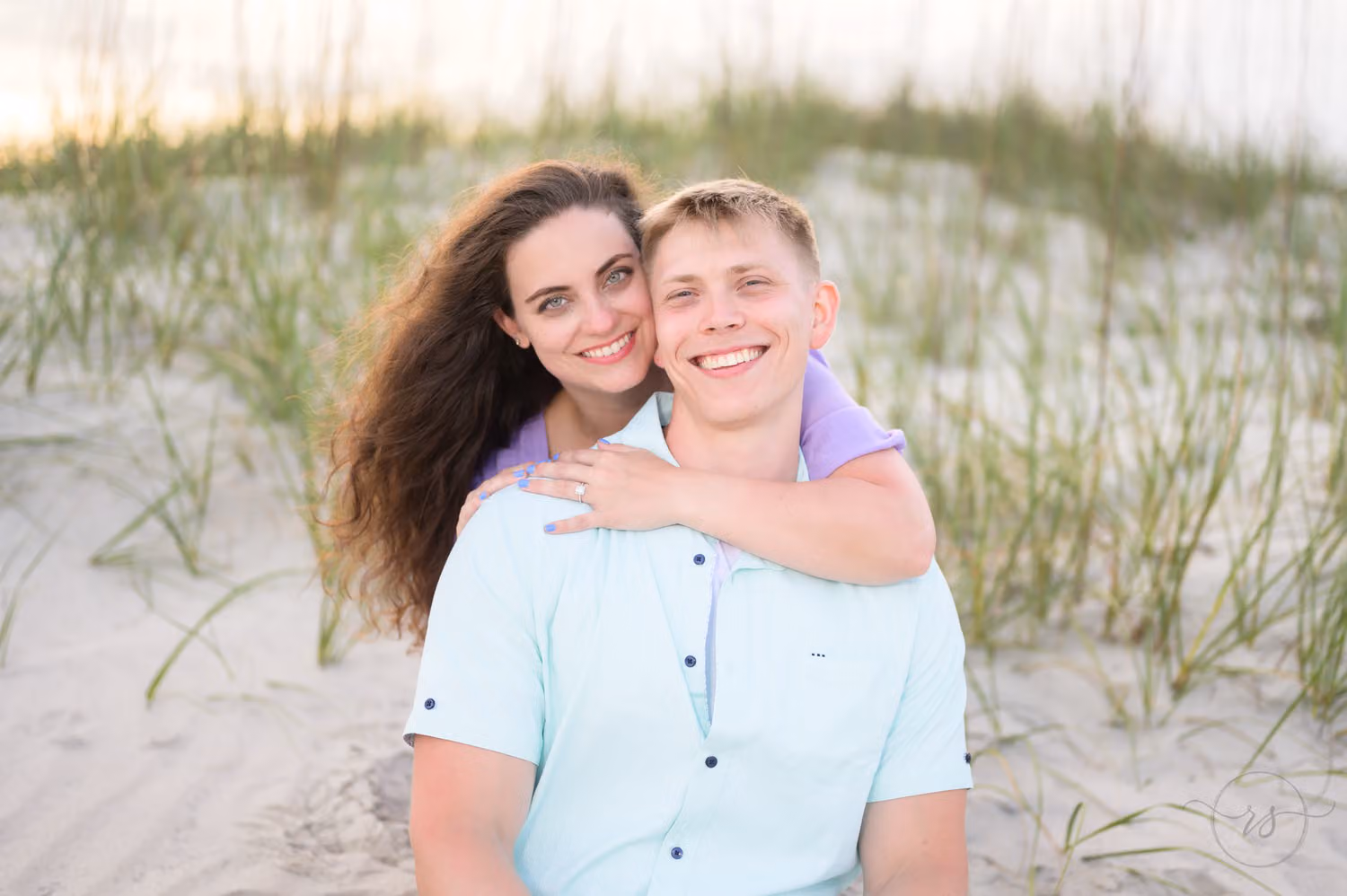 Woman hugging her fiancé from behind as they sit in the sand among the sea oats at Huntington Beach State Park