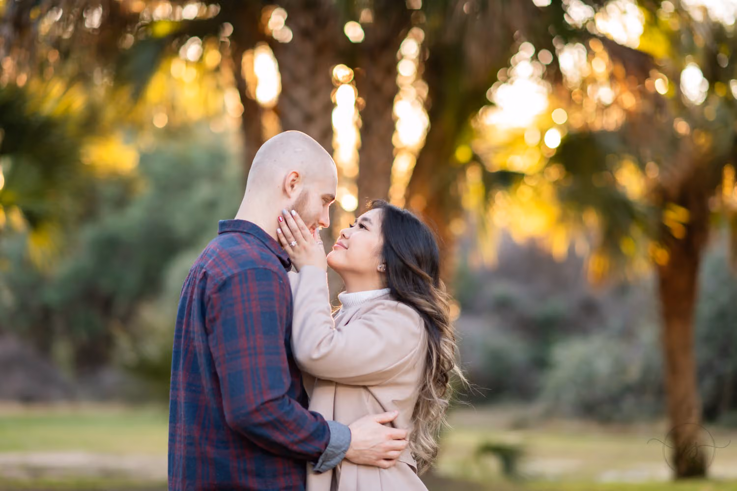Couple standing close with warm sunlight glowing through the palm trees behind them at Huntington Beach State Park