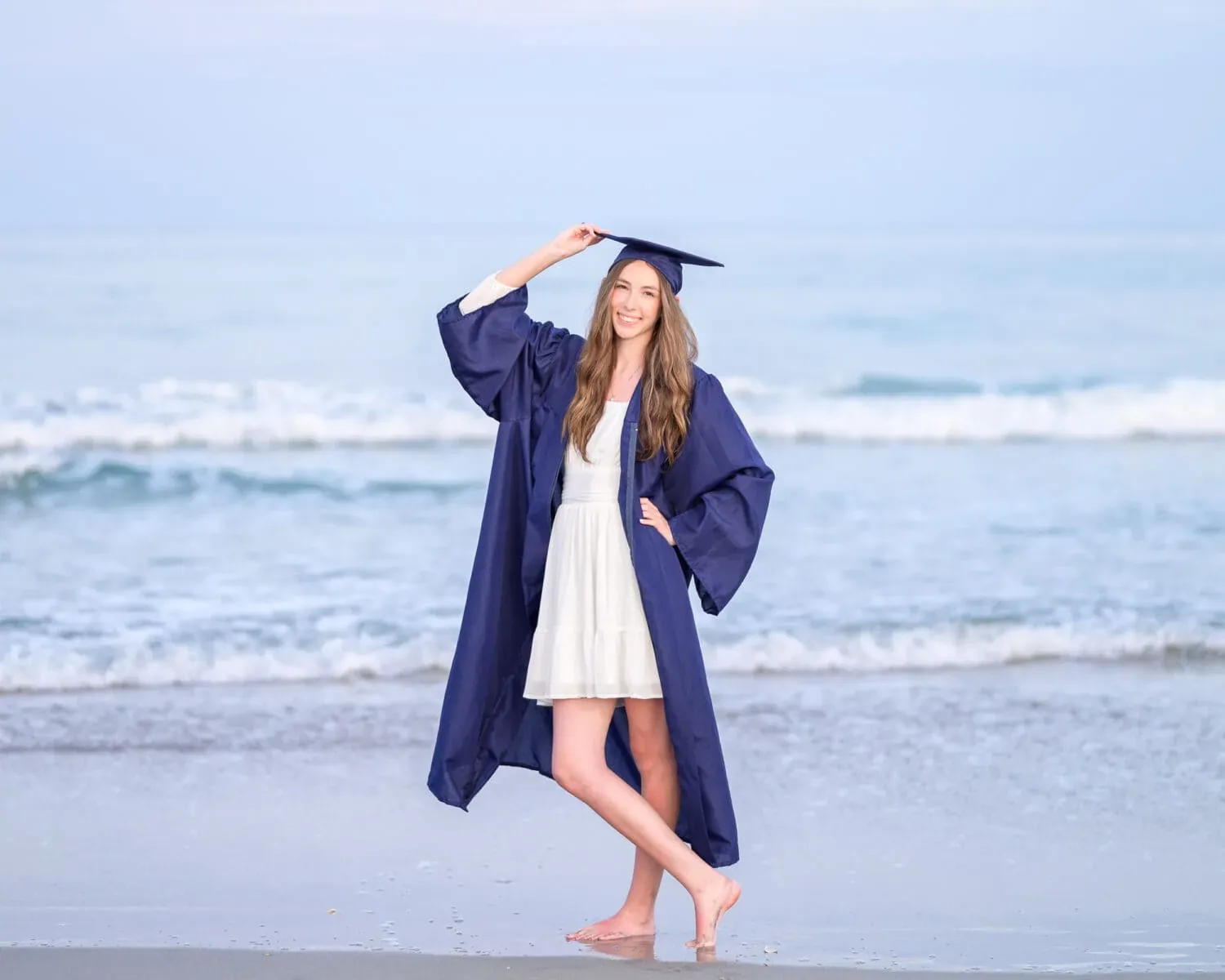 Graduate in a navy gown posing barefoot with one hand on her cap by the waves at Huntington Beach State Park