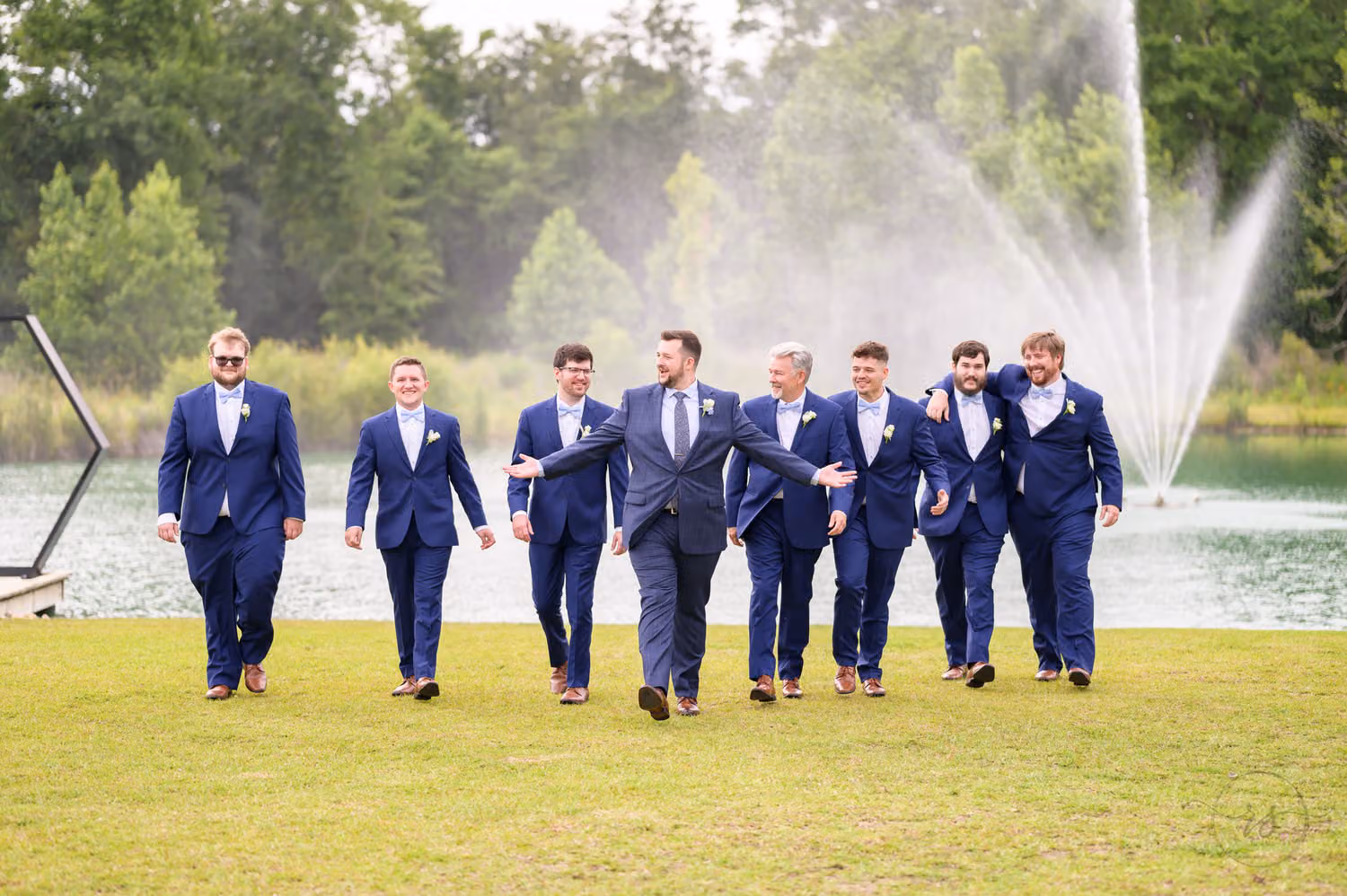 Groom and groomsmen in navy suits walking across the lawn in front of a lake fountain at The Venue at White Oaks Farm
