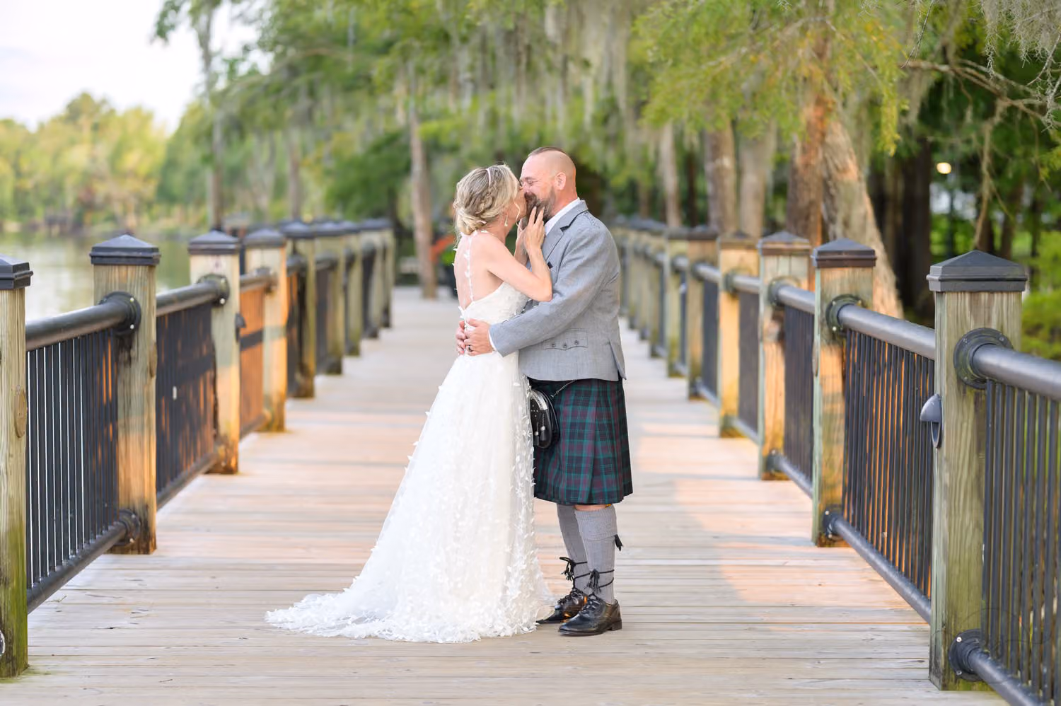 Bride and groom in a gray jacket and tartan kilt kissing on the wooden boardwalk under mossy trees at the Conway Riverwalk