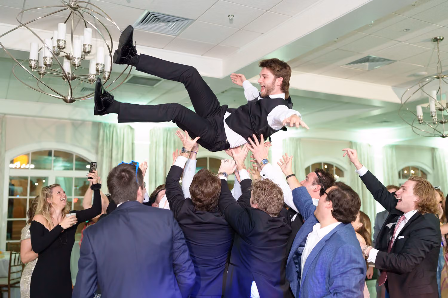 Guests tossing the groom high into the air under the chandeliers on the dance floor at 21 Main Events