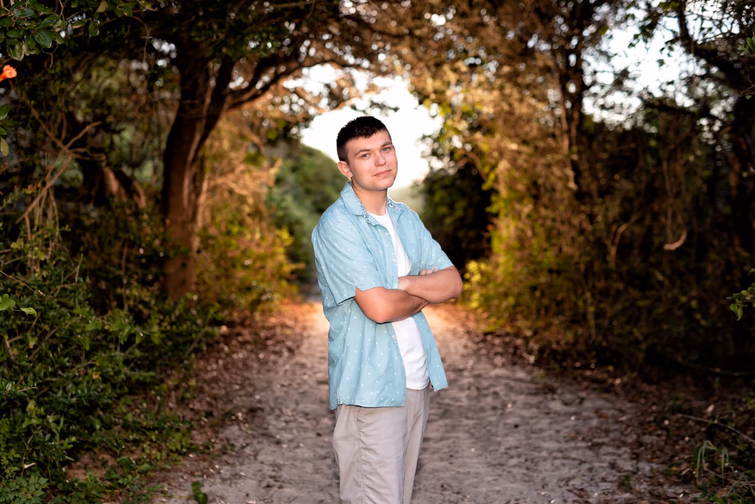 Senior guy in a teal shirt with arms crossed on a dark tree-lined path, backlit by strobe at Huntington Beach State Park