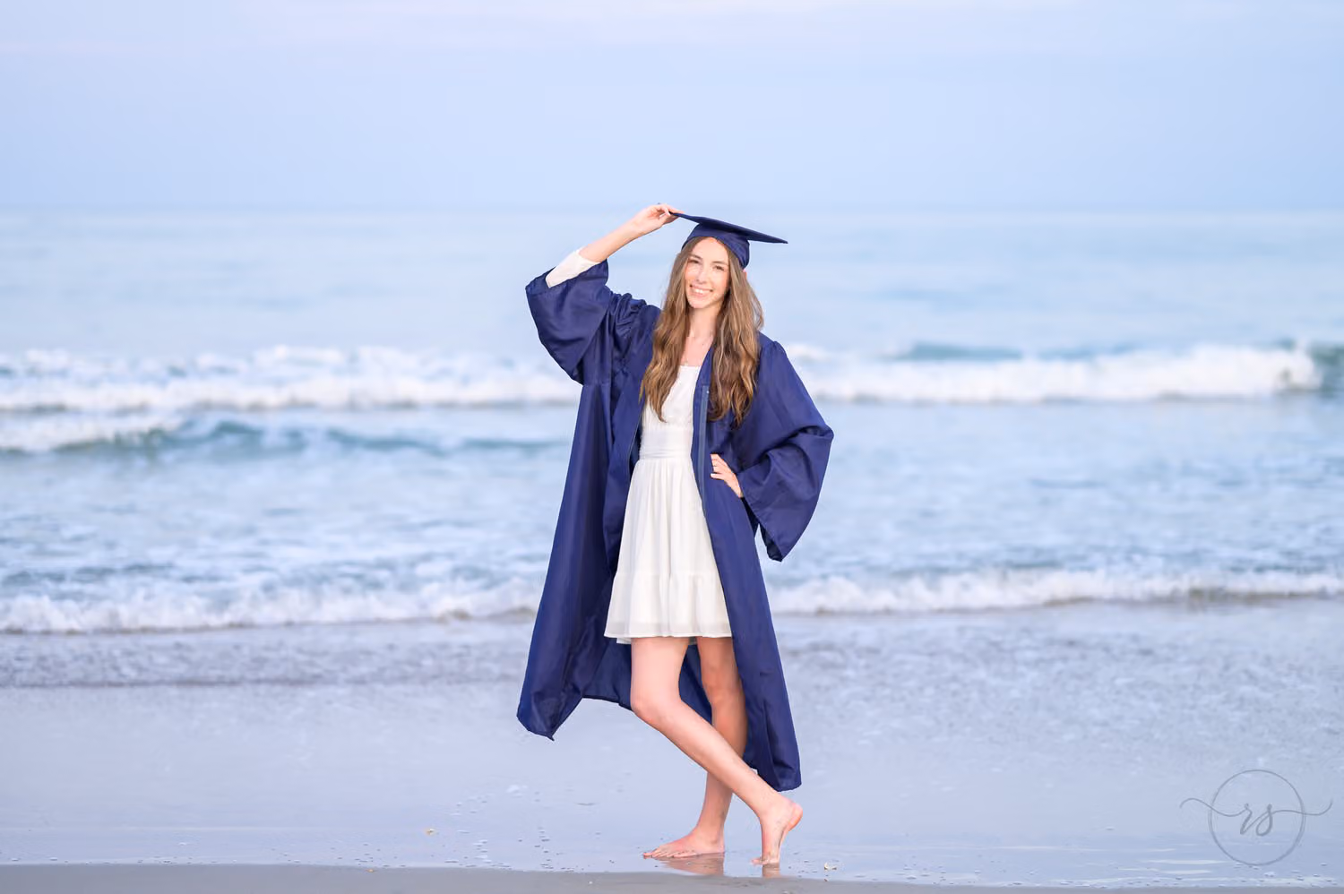 Graduate in a navy gown and white dress posing barefoot with one hand on her cap by the waves at Huntington Beach State Park
