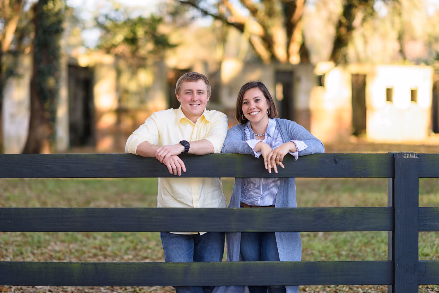 Engaged couple leaning on a dark wooden fence with the old hotel and oak trees behind them in Pawleys Island