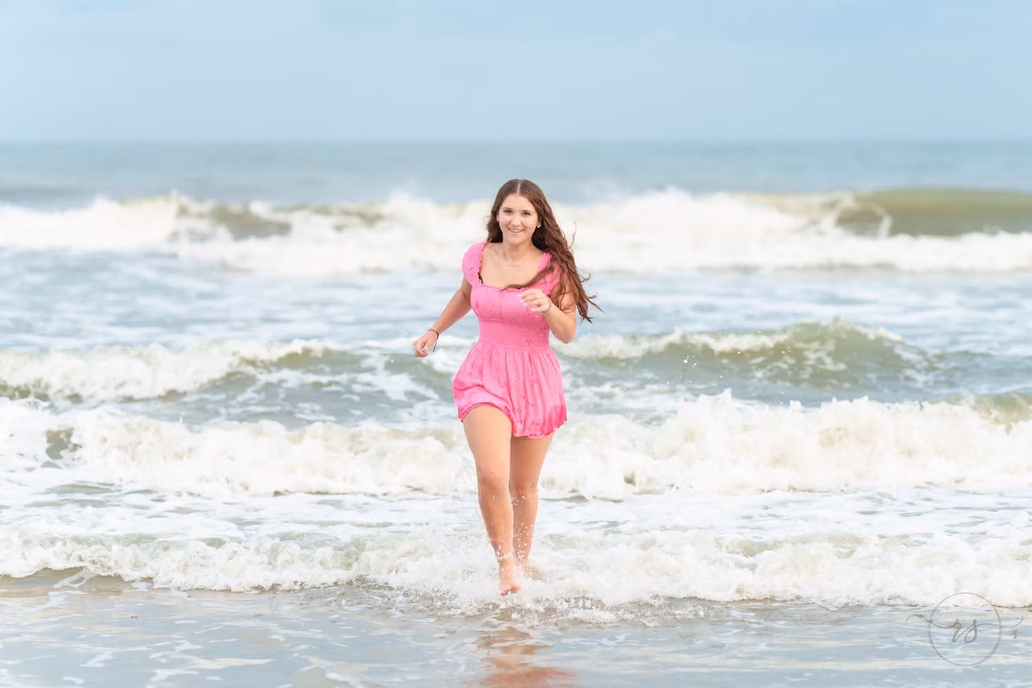 Senior in a pink romper running and laughing through the breaking waves at Huntington Beach State Park