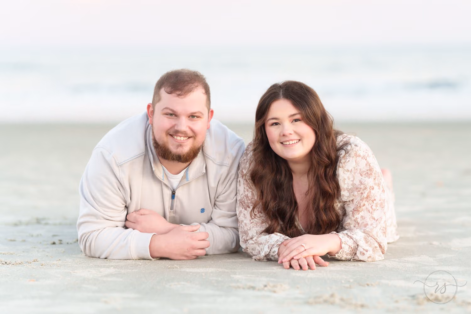 Newly engaged couple lying side by side on the sand, smiling at the camera, at Huntington Beach State Park