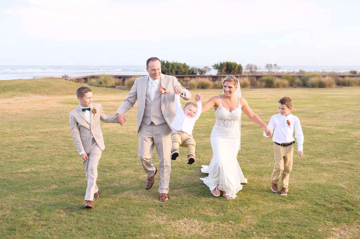 Newlyweds swinging a toddler as two boys run beside them on the oceanfront lawn at Dunes Golf & Beach Club