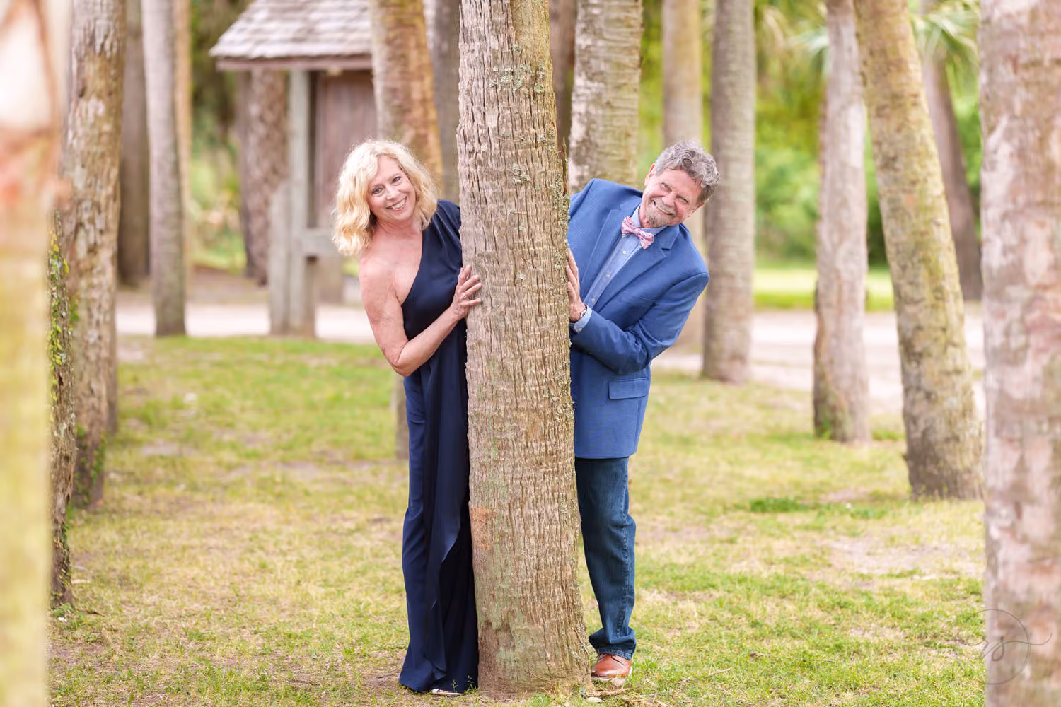Smiling older couple peeking out from behind palm tree trunks on a grassy lawn at Huntington Beach State Park