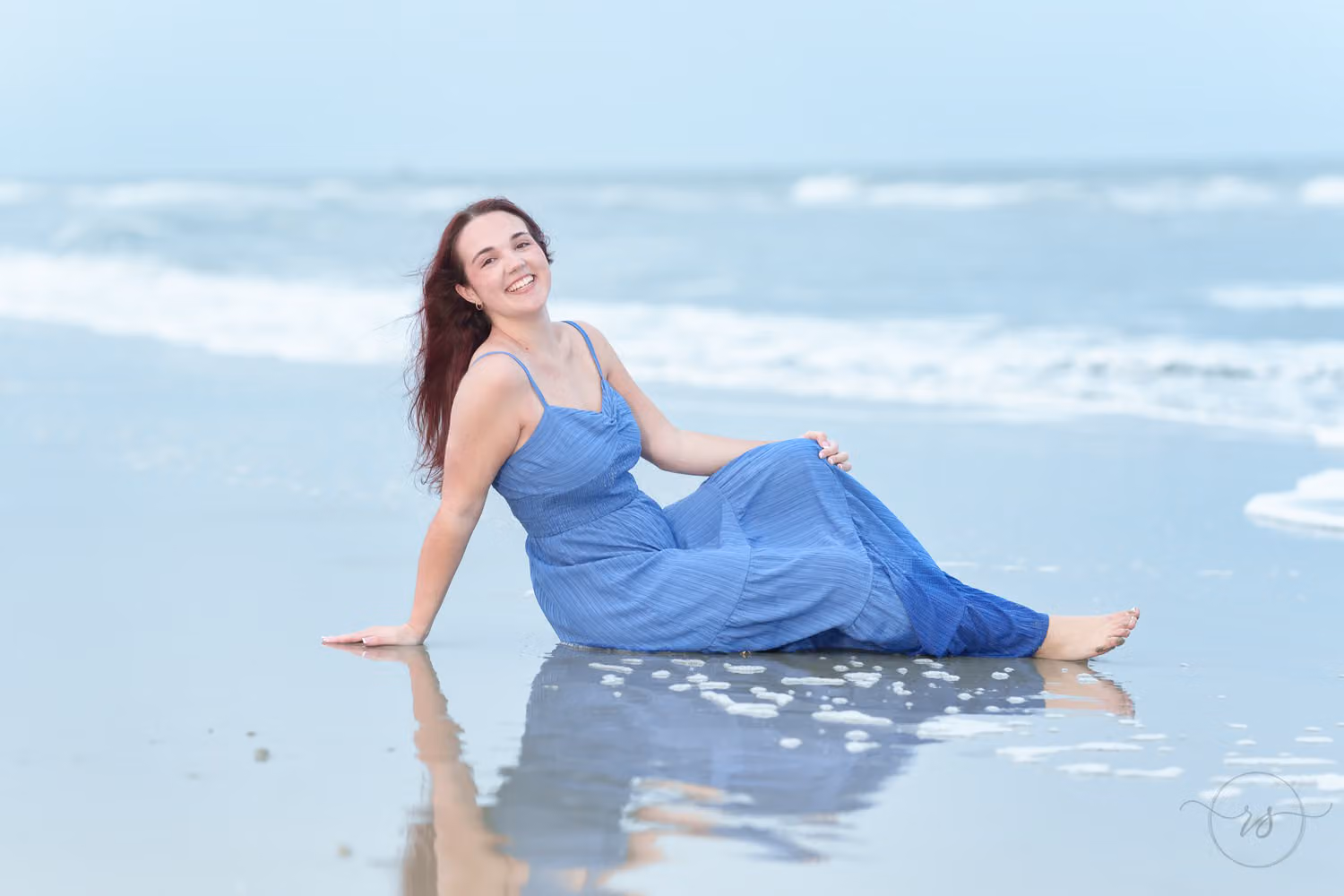 Senior in a blue dress sitting at the water's edge with her reflection in the wet sand at Huntington Beach State Park