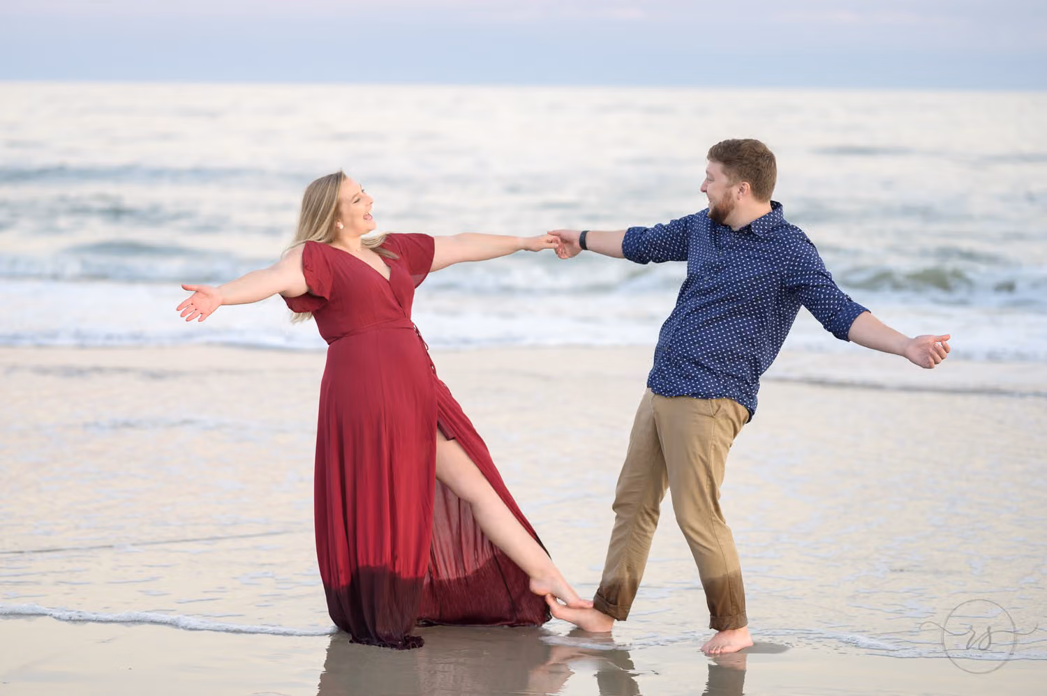 Couple holding hands and leaning apart playfully in the shallow water, she in a red dress, at Huntington Beach State Park