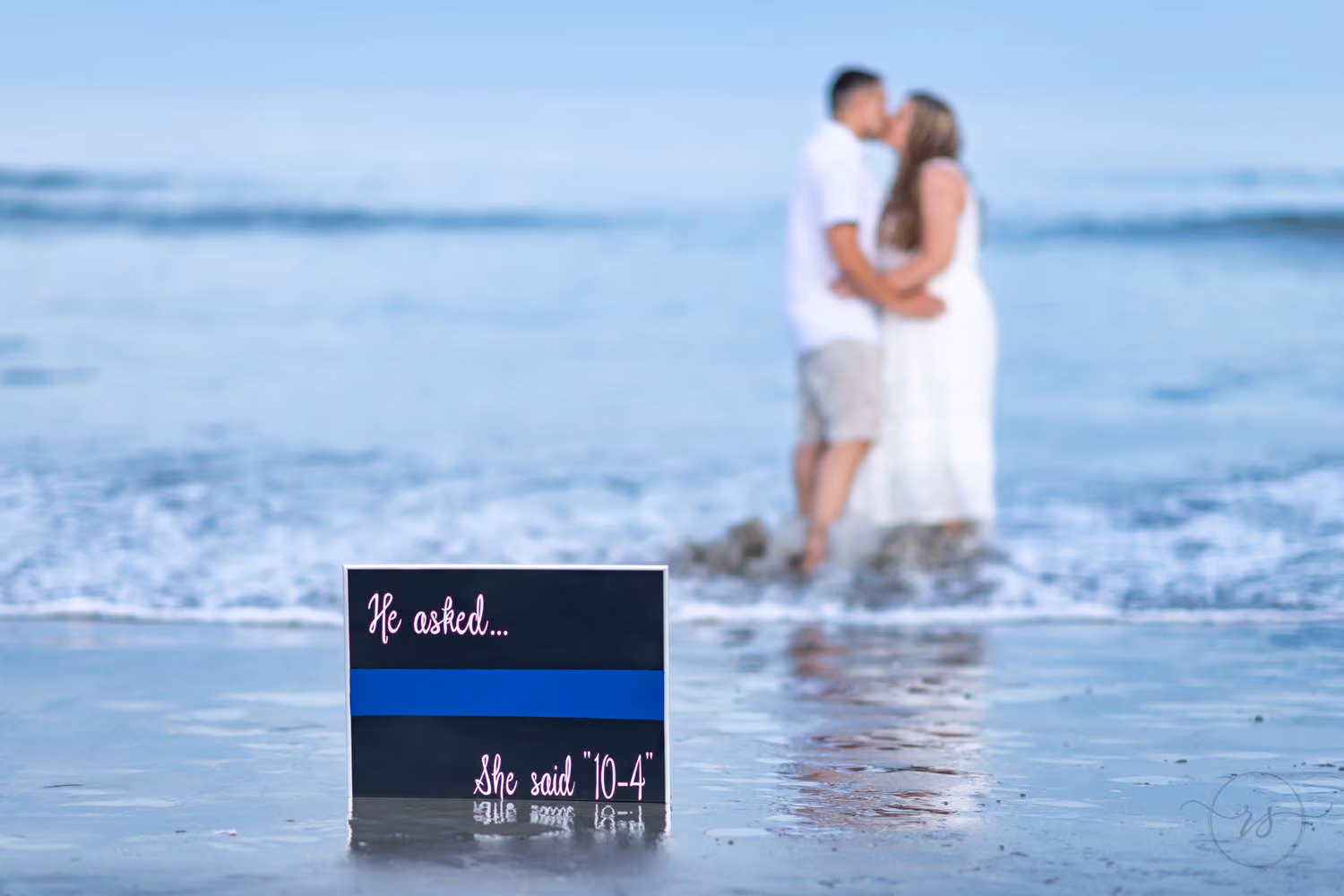 Couple kissing in the surf behind a thin blue line sign reading he asked, she said 10-4 at Huntington Beach State Park