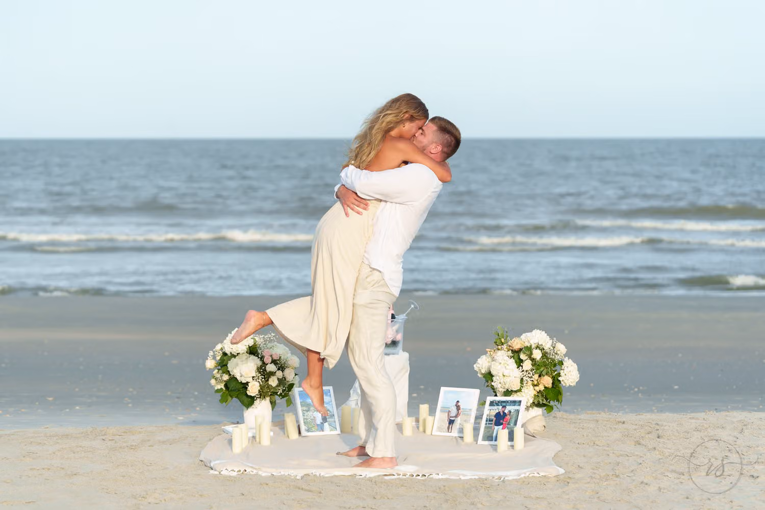 Newly engaged man lifting his fiancée beside a beach proposal setup with flowers and candles in Pawleys Island
