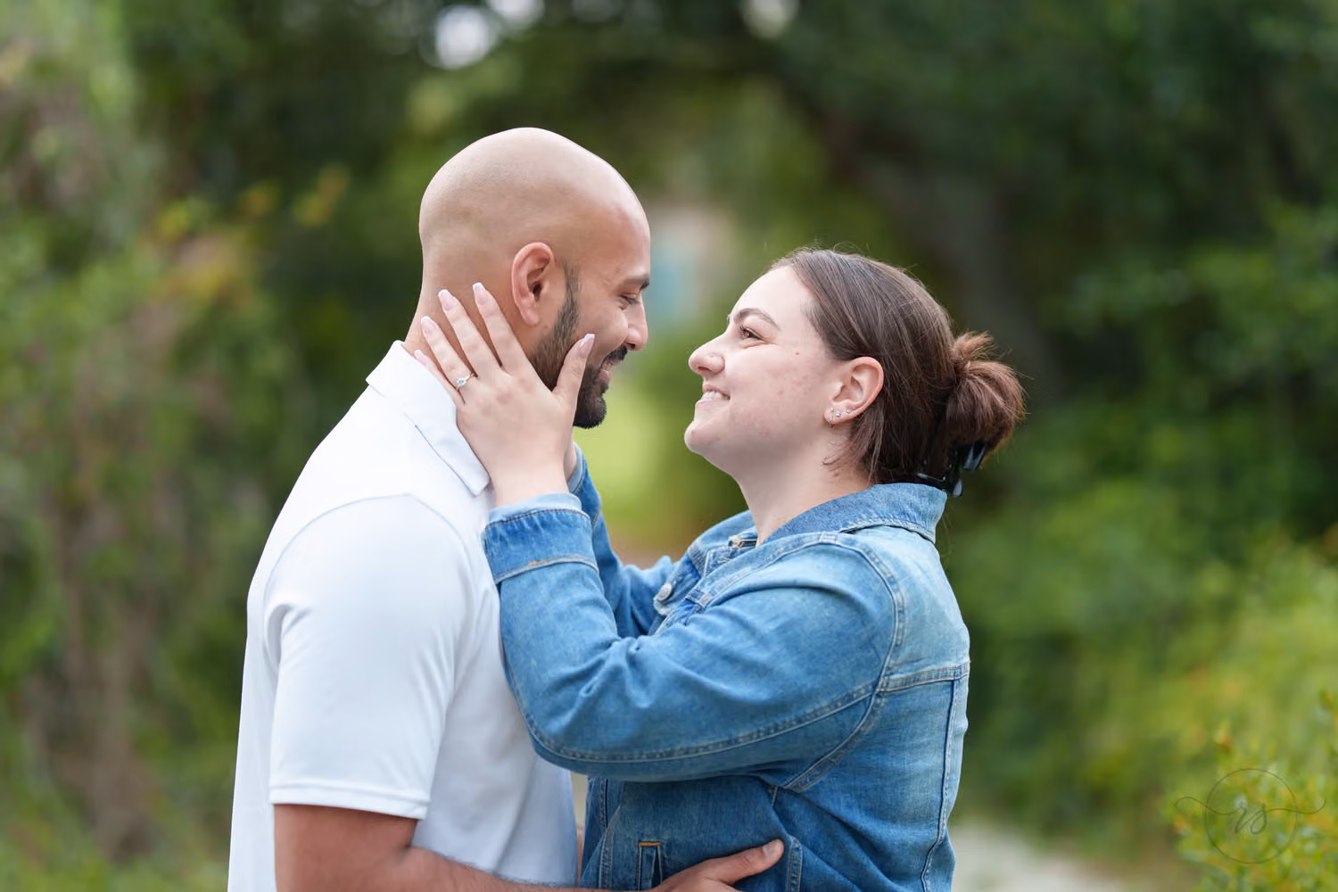 Woman in a denim jacket holding her fiancé's face as they smile at each other under the trees at Huntington Beach State Park