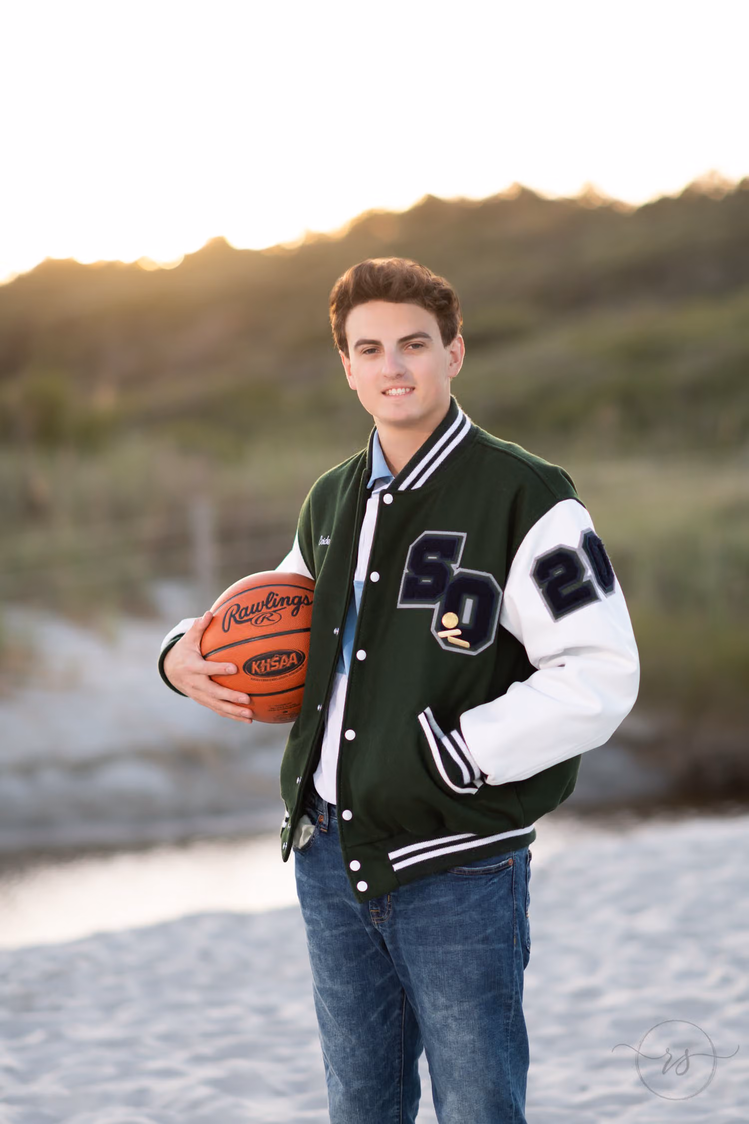Senior in a green and white letterman jacket holding a basketball in front of the dunes at Myrtle Beach State Park